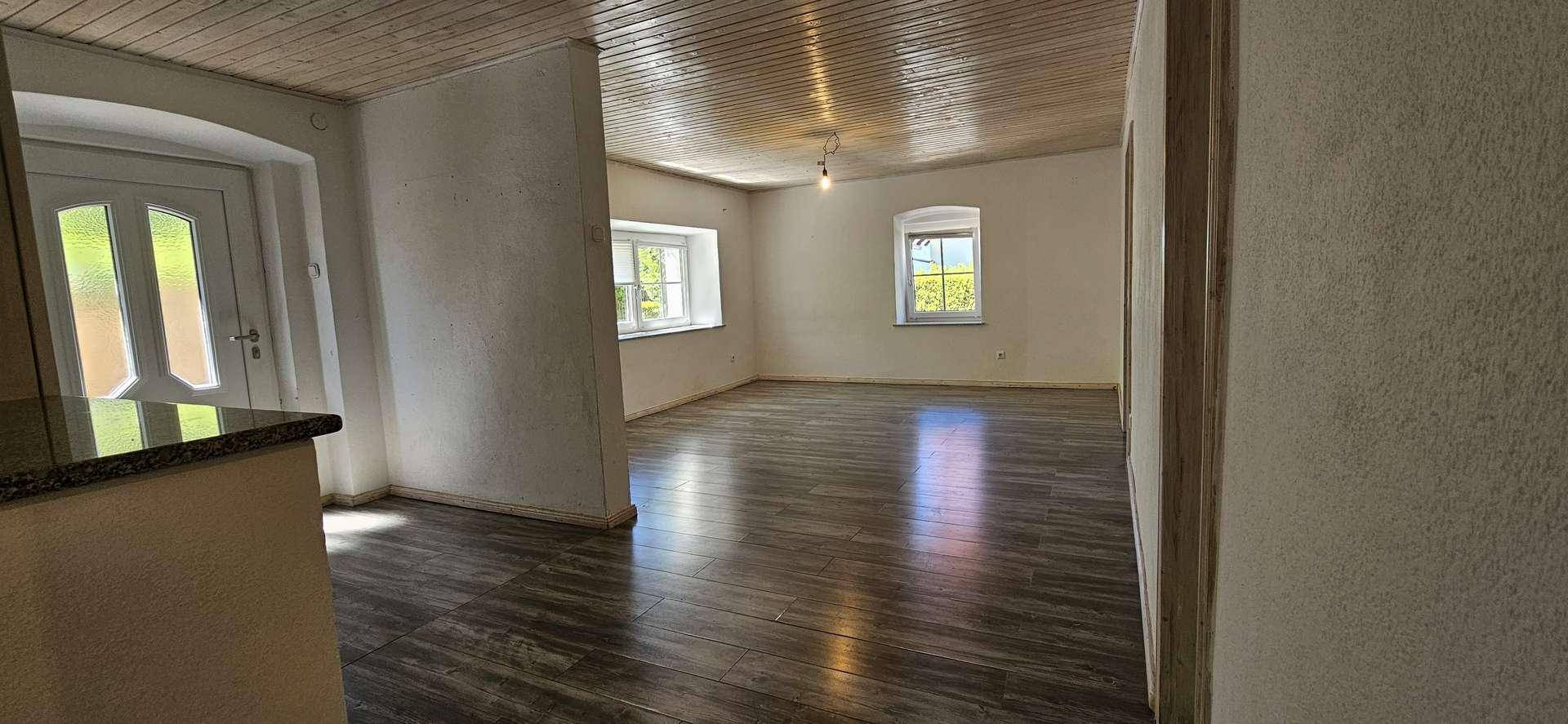 Geräumiger Wohnraum mit Holzdielen-Decke, dunklem Laminatboden und mehreren Fenstern für viel Tageslicht.