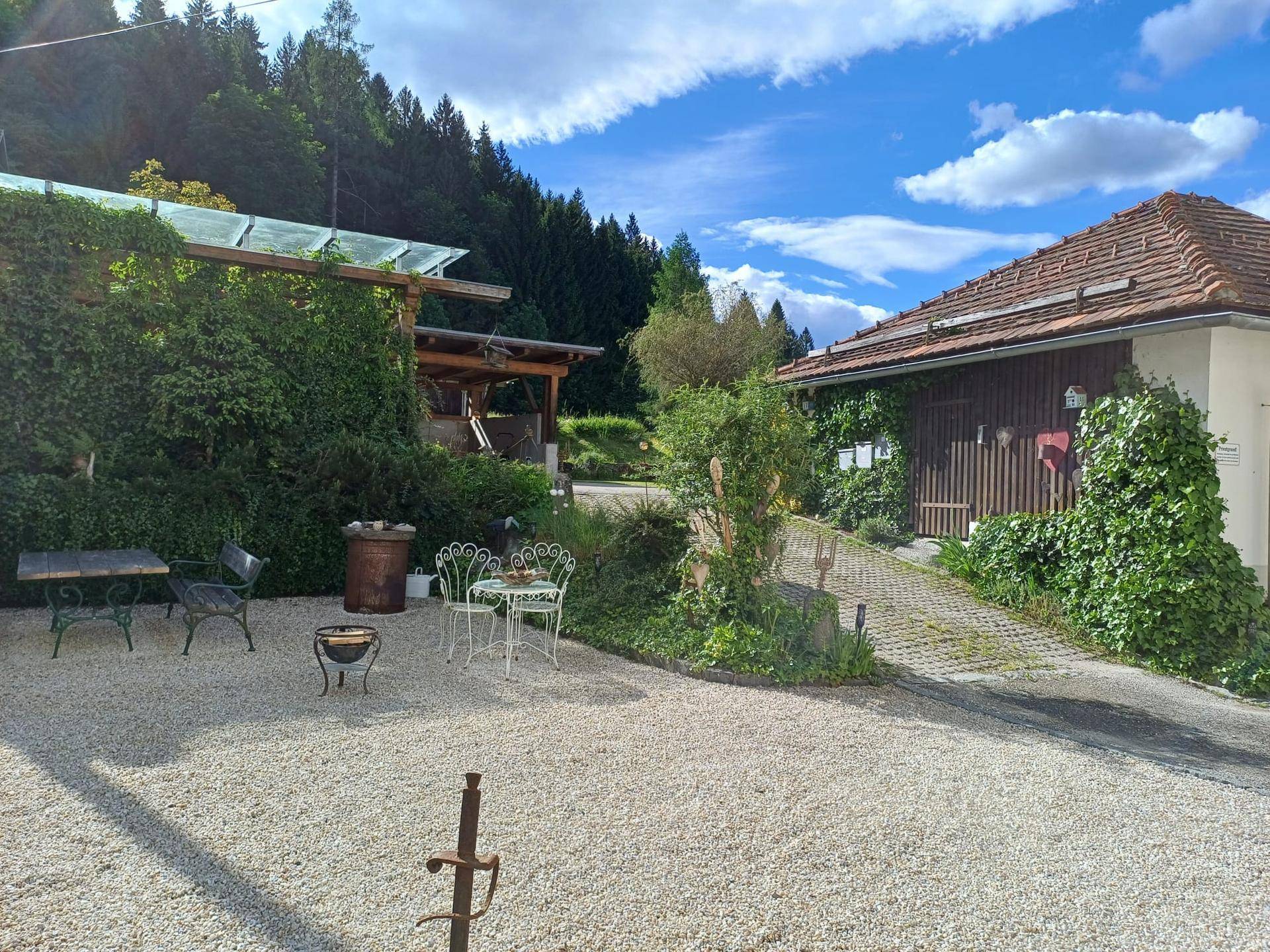 Außenbereich mit Kiesfläche, viel Grün und einem Nebengebäude im Hintergrund.