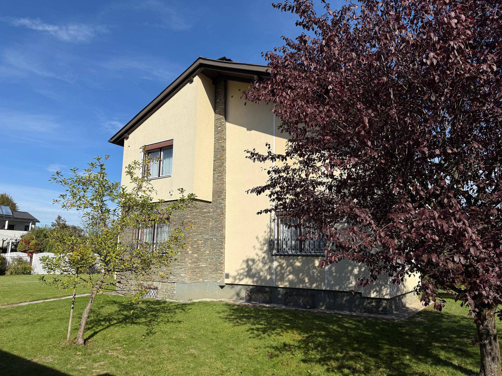 Seitenansicht des Hauses mit heller Fassade, Steinelementen und einem gepflegten Vorgarten unter blauem Himmel.