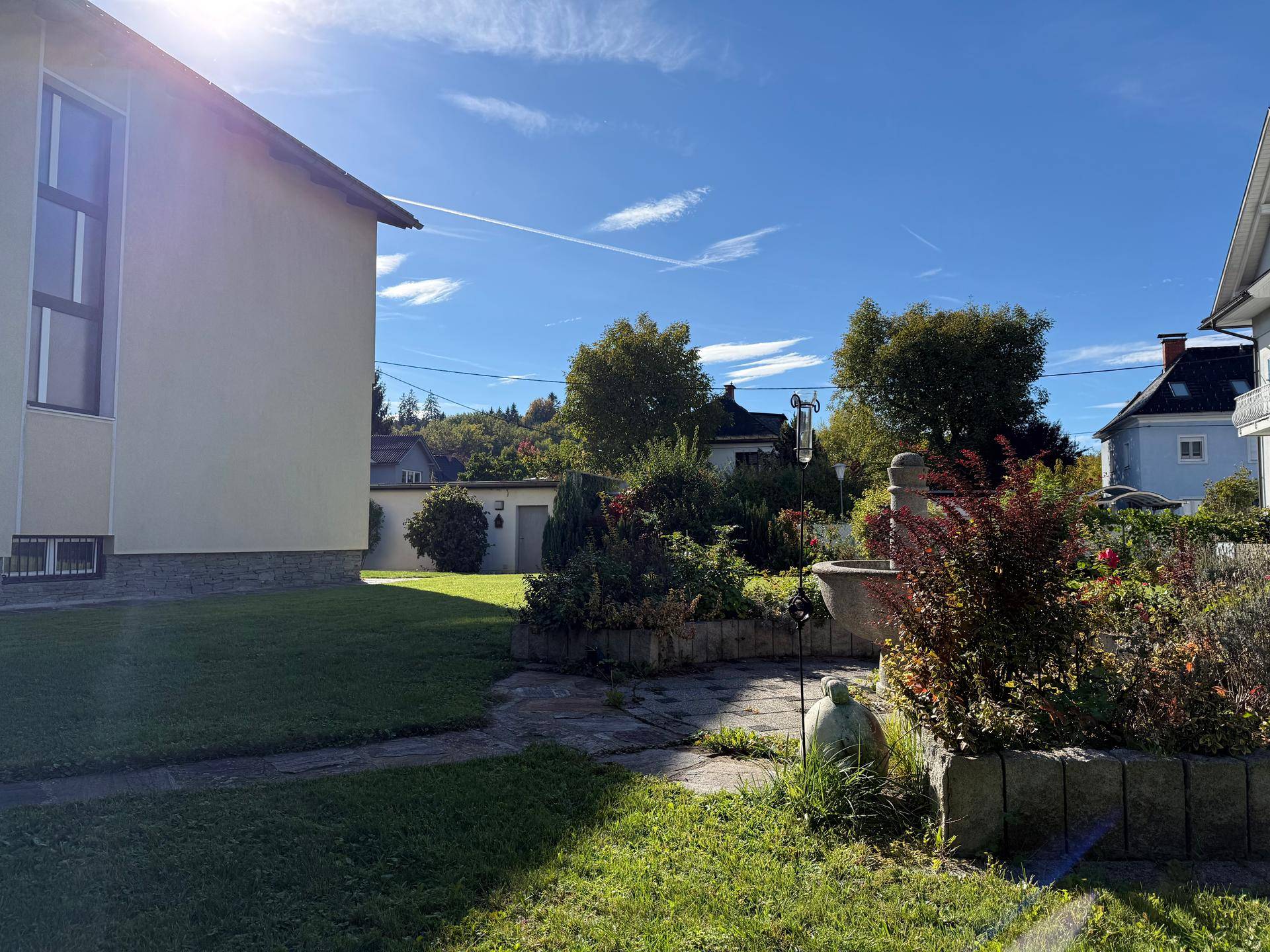 Gartenbereich mit dekorativem Brunnen, Blumenbeeten und gepflegten Pflanzen unter blauem Himmel.
