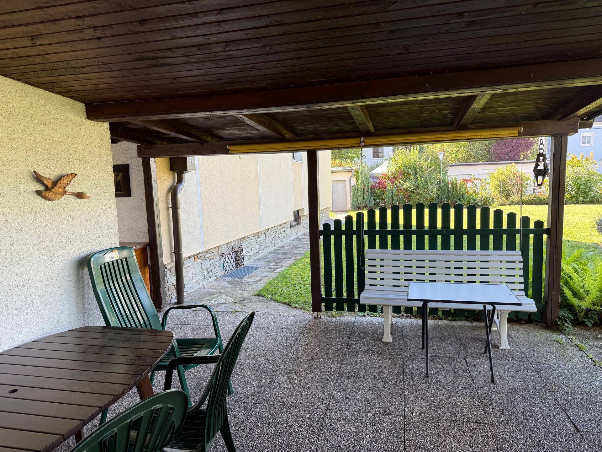 Überdachte Terrasse mit Holzdach, Gartenmöbeln und einer weißen Bank, ideal für entspannte Stunden im Freien.