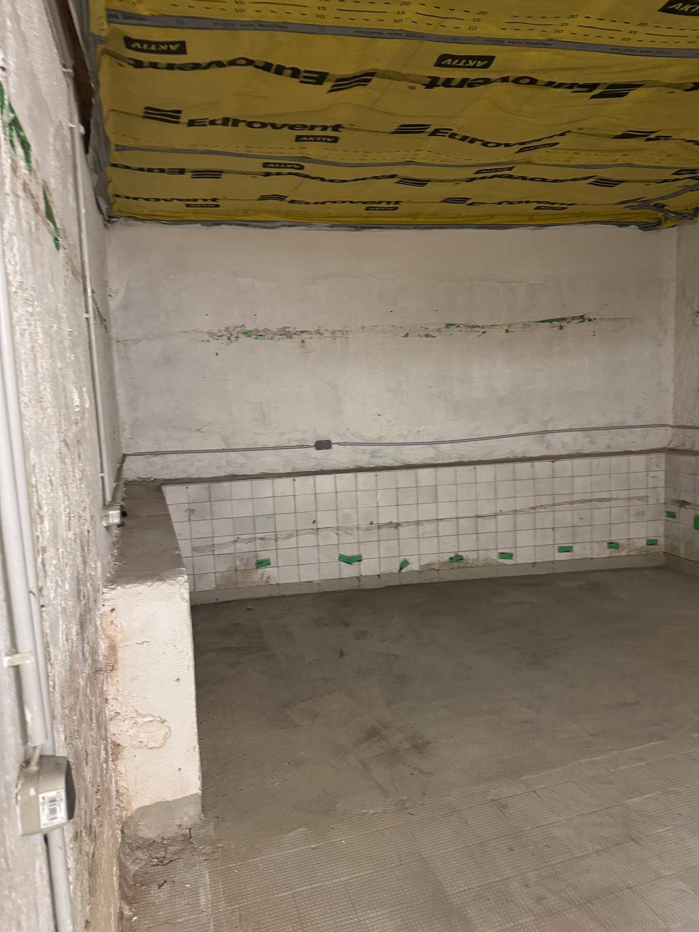 Raum im Rohbau mit gefliesten Wänden im unteren Bereich und sichtbarer Isolierung an der Decke.