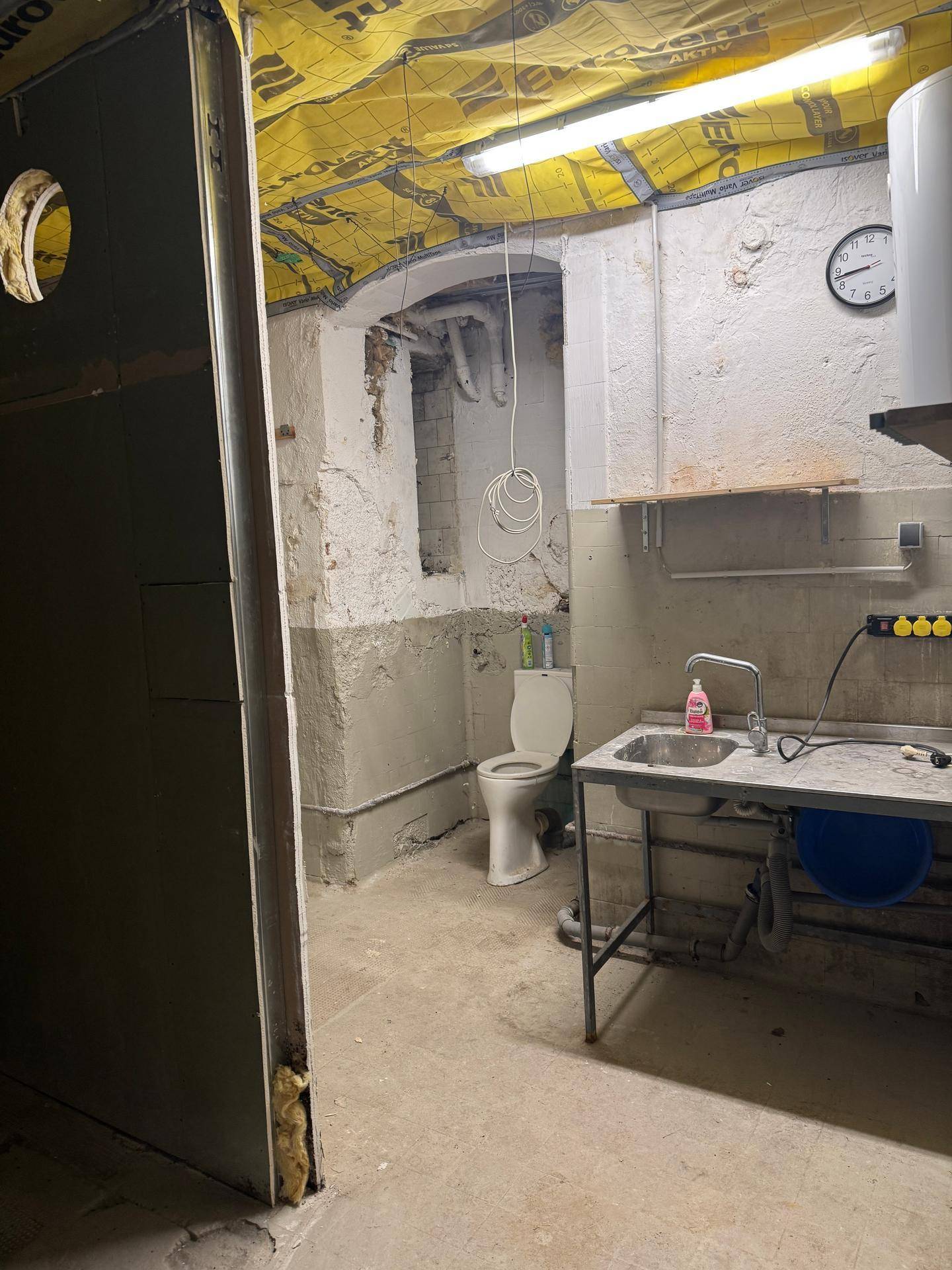 Einfacher Hauswirtschaftsraum im Keller mit Toilette, Waschbecken und Warmwasserbereiter in rohem Zustand.