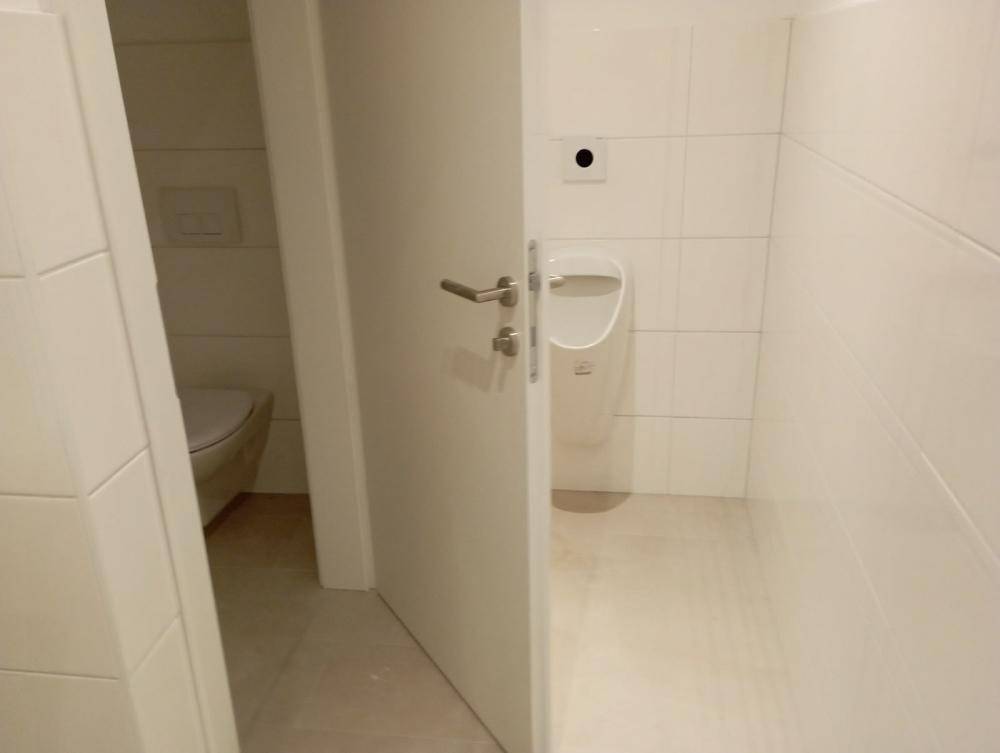 Gäste-WC mit Urinal und Toilette, getrennt durch eine Tür, in weiß gefliestem Raum.