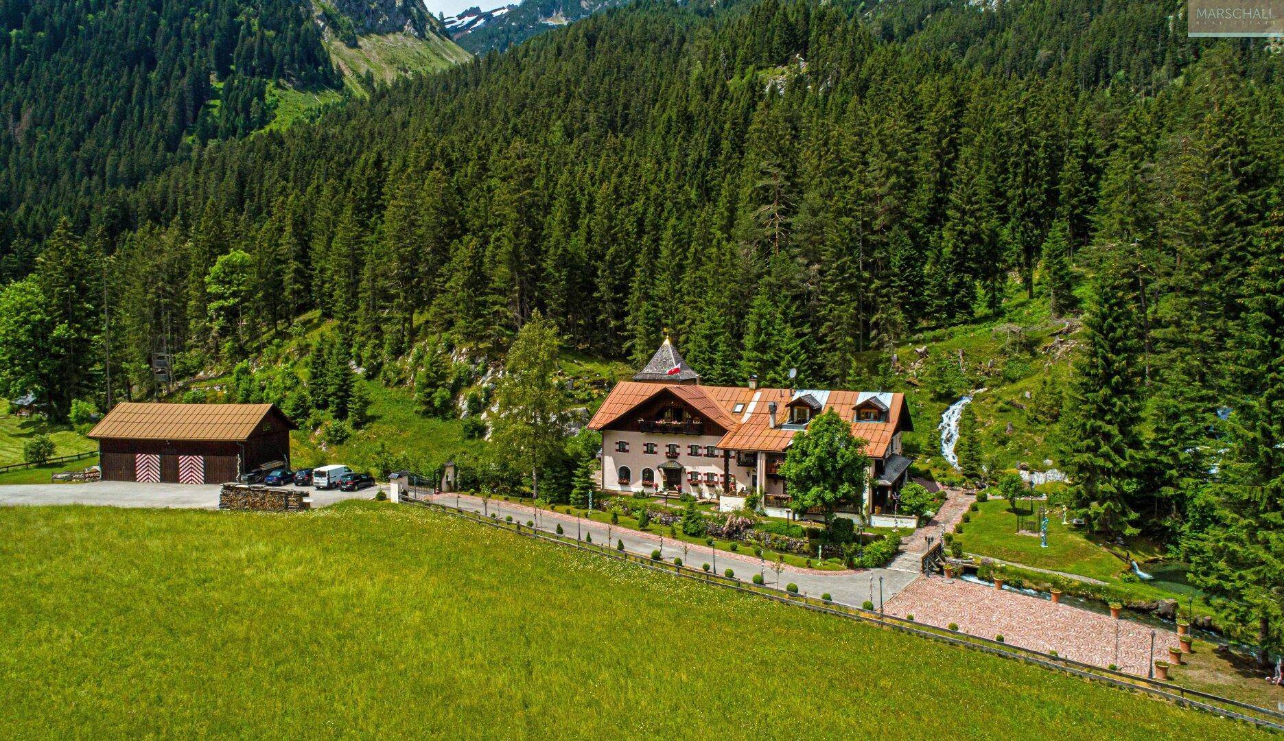Luftaufnahme eines Anwesens im Grünen, umgeben von Wäldern und Bergen, mit Zufahrt und Nebengebäude.