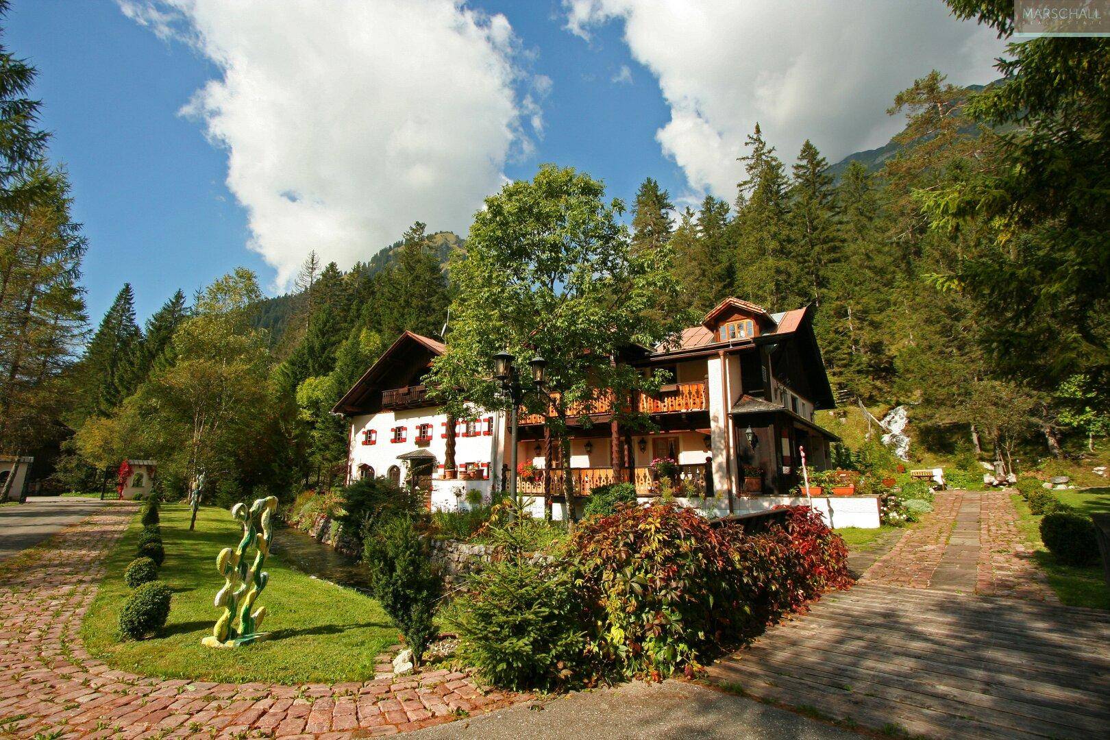 Charmantes Alpenhaus mit Balkonen und gepflegtem Garten, umgeben von hohen Bäumen und Bergen.