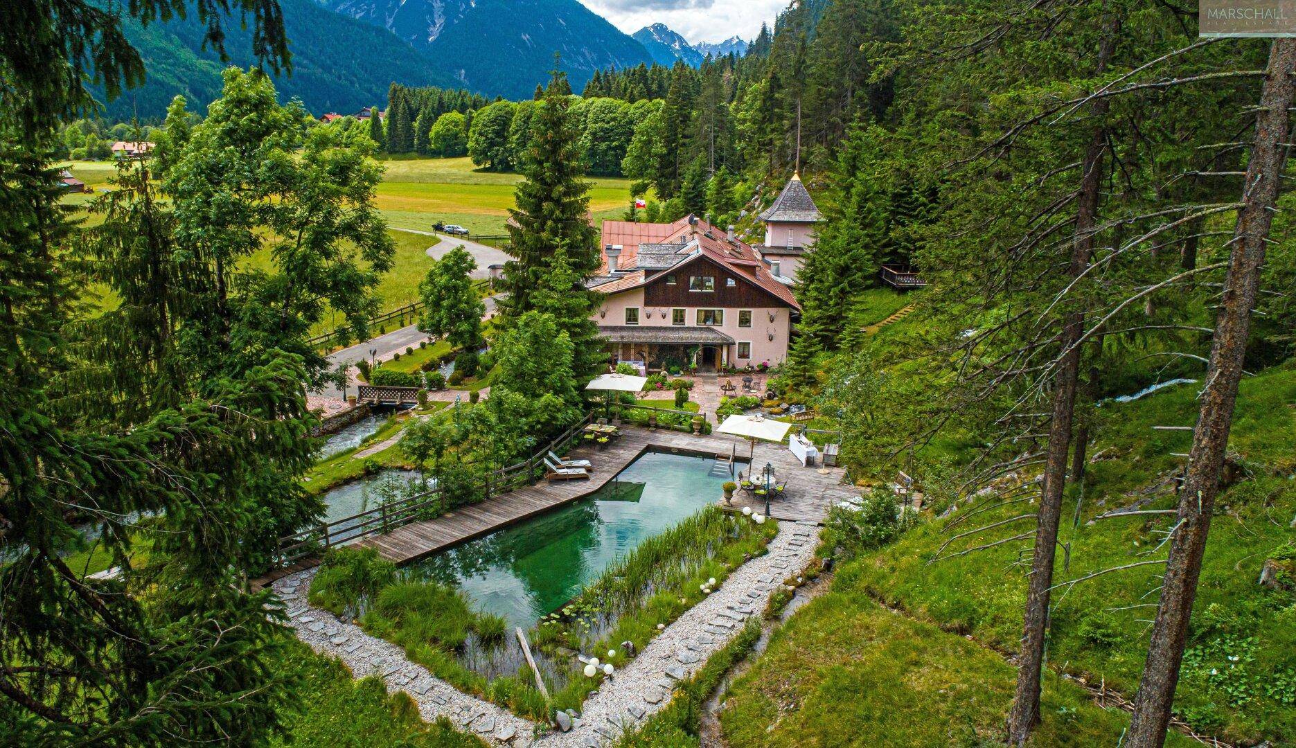 Panoramablick auf das Anwesen mit Schwimmteich, Gartenwegen und umliegenden Wäldern und Bergen.