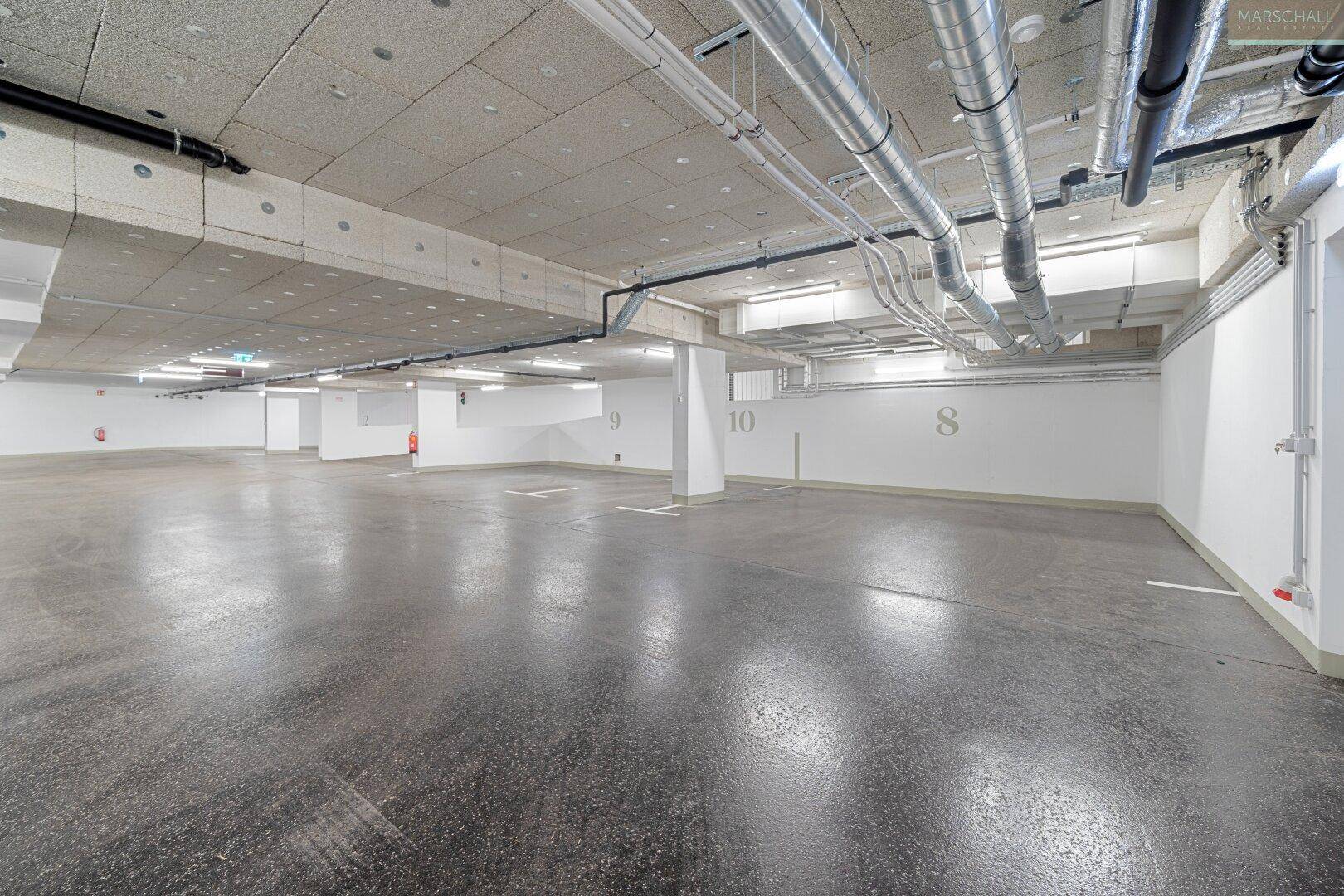 Geräumige Tiefgarage mit vielen Stellplätzen, sichtbaren Installationen und Betonboden.