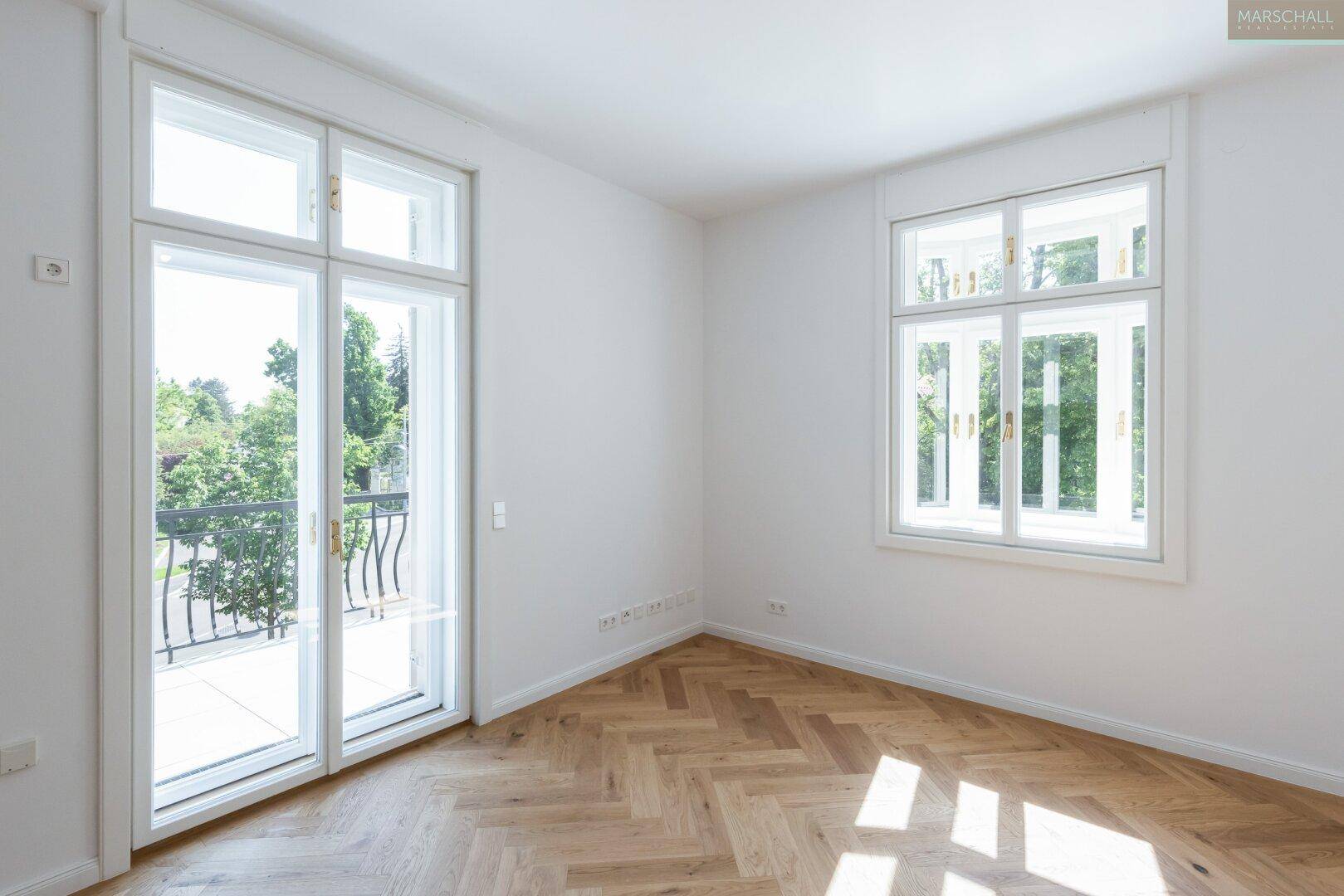 Lichtdurchfluteter Raum mit Fischgrätparkett, großen Fenstern und Zugang zu einem Balkon.