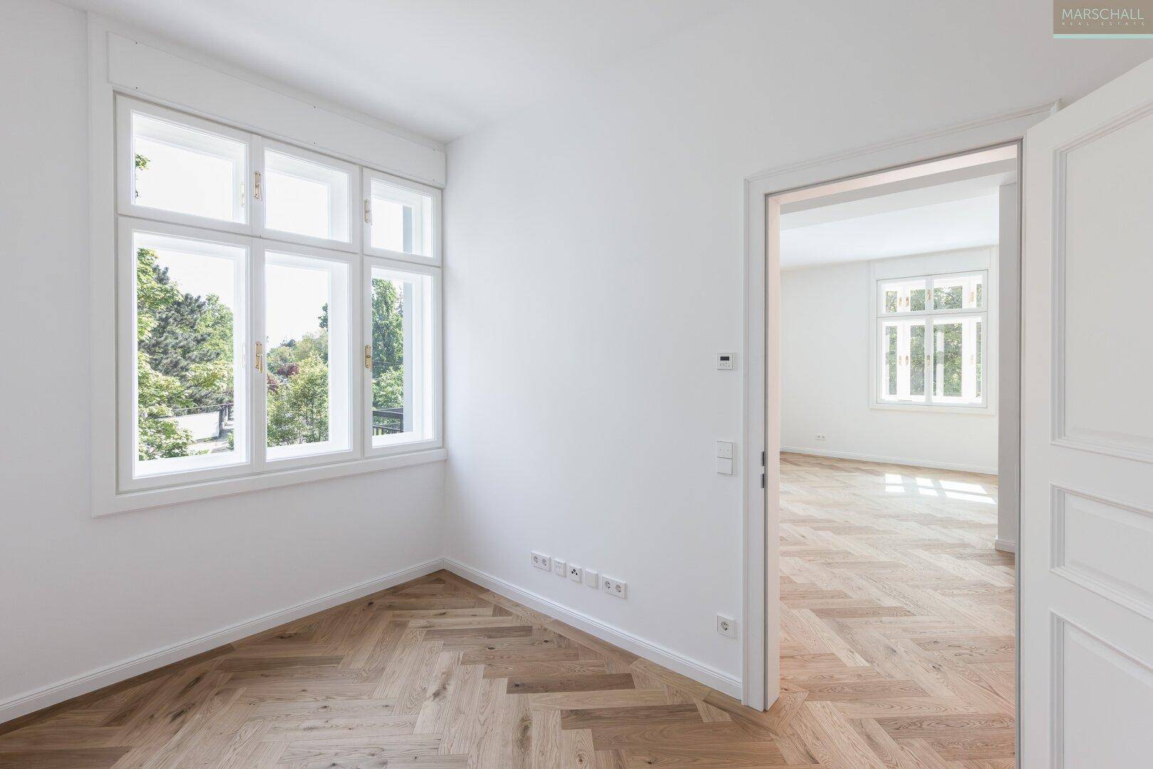 Helles Zimmer mit Fischgrätparkett und großen Fenstern, die einen Blick ins Grüne bieten.