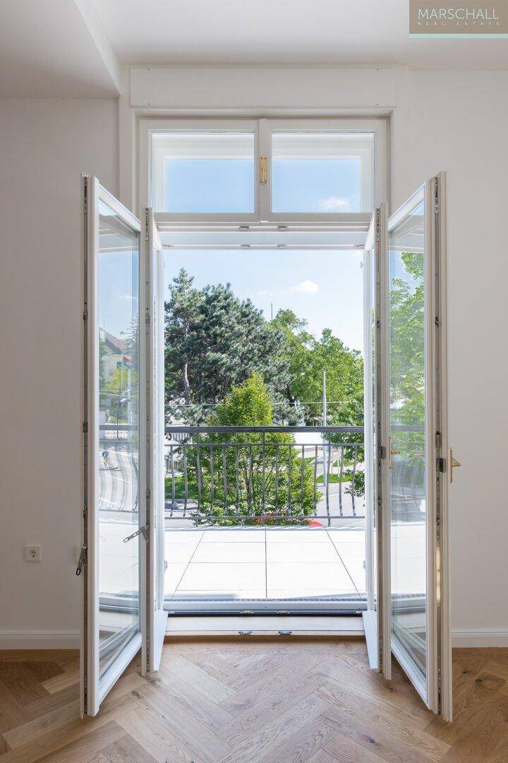 Heller Raum mit geöffneten Fenstertüren, die den Blick auf einen Balkon und grüne Bäume freigeben.