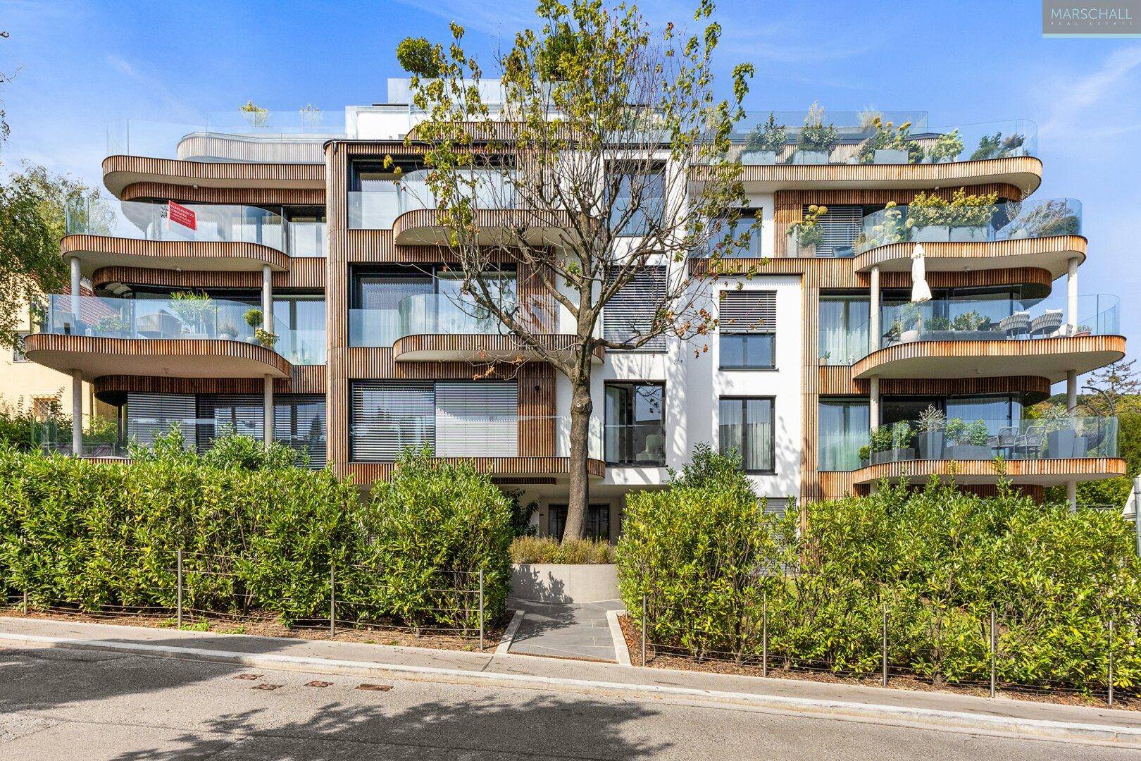 Moderne Wohnhausfassade mit geschwungenen Balkonen, großen Fenstern und grünen Hecken.