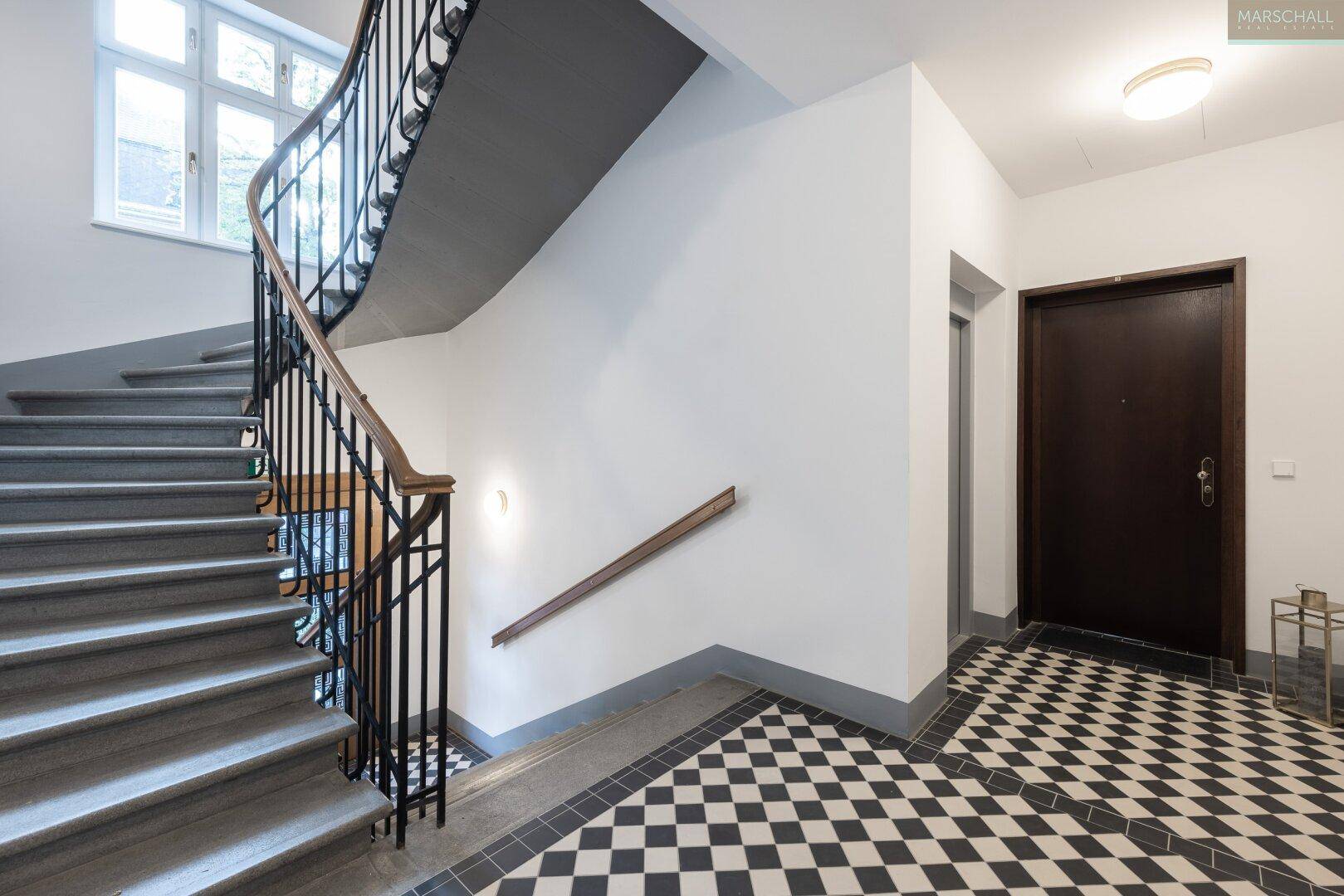 Elegantes Treppenhaus mit geschwungener Treppe, schmiedeeisernem Geländer und Schachbrettmuster-Fliesenboden.
