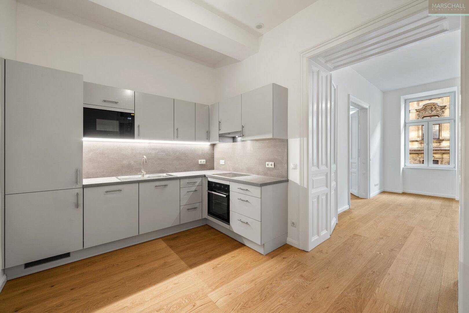 Moderne Einbauküche in Grau mit integrierten Geräten und hellem Parkettboden, offen zum Wohnbereich.
