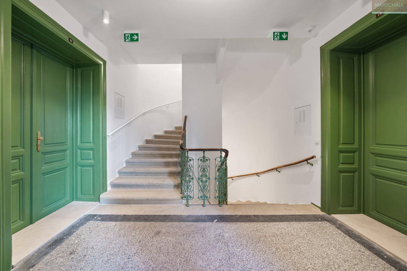 Historisches Treppenhaus mit geschwungener Steintreppe, schmiedeeisernem Geländer und auffälligen grünen Türen.