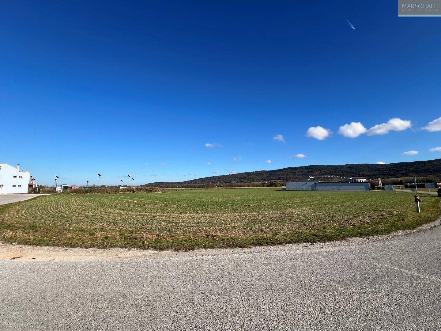 Offenes Feld mit einer Straße im Vordergrund, umgeben von Gewerbegebäuden und einer Bergkette am Horizont.