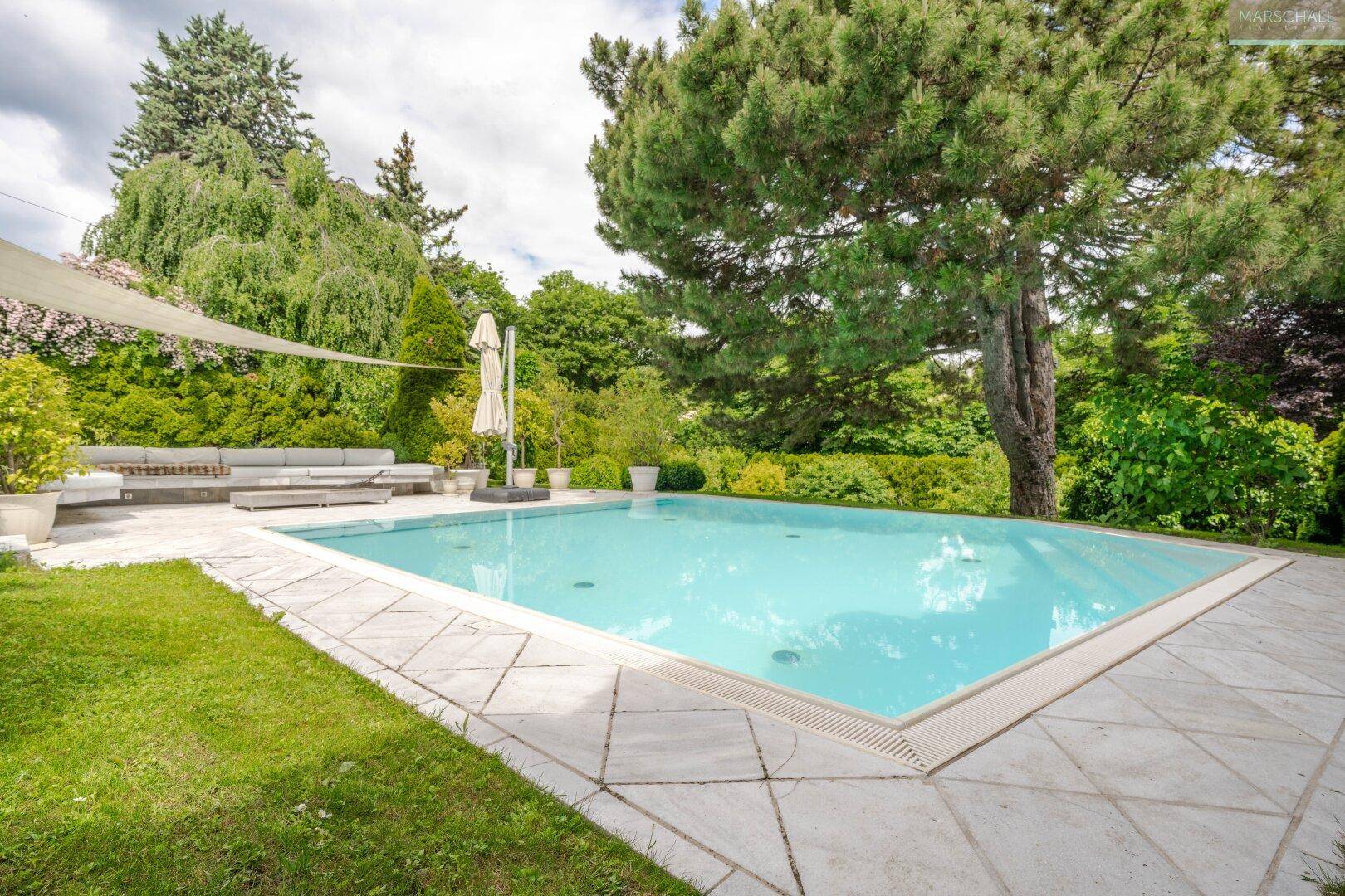 Gepflegter Swimmingpool-Bereich im Garten mit Steinplatten, Liegemöglichkeiten und umgebenden Bäumen.