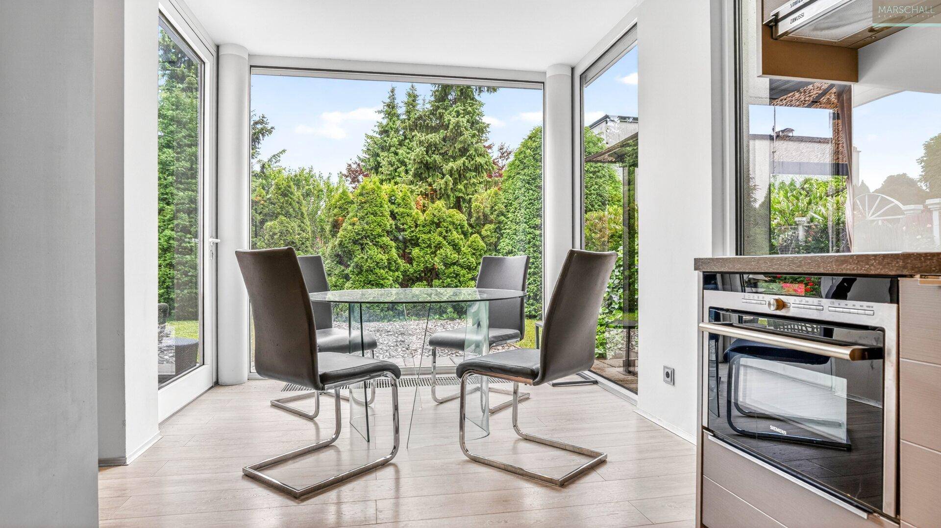 Lichtdurchfluteter Essbereich mit modernem Glastisch und Stühlen, umgeben von üppigem Grün.