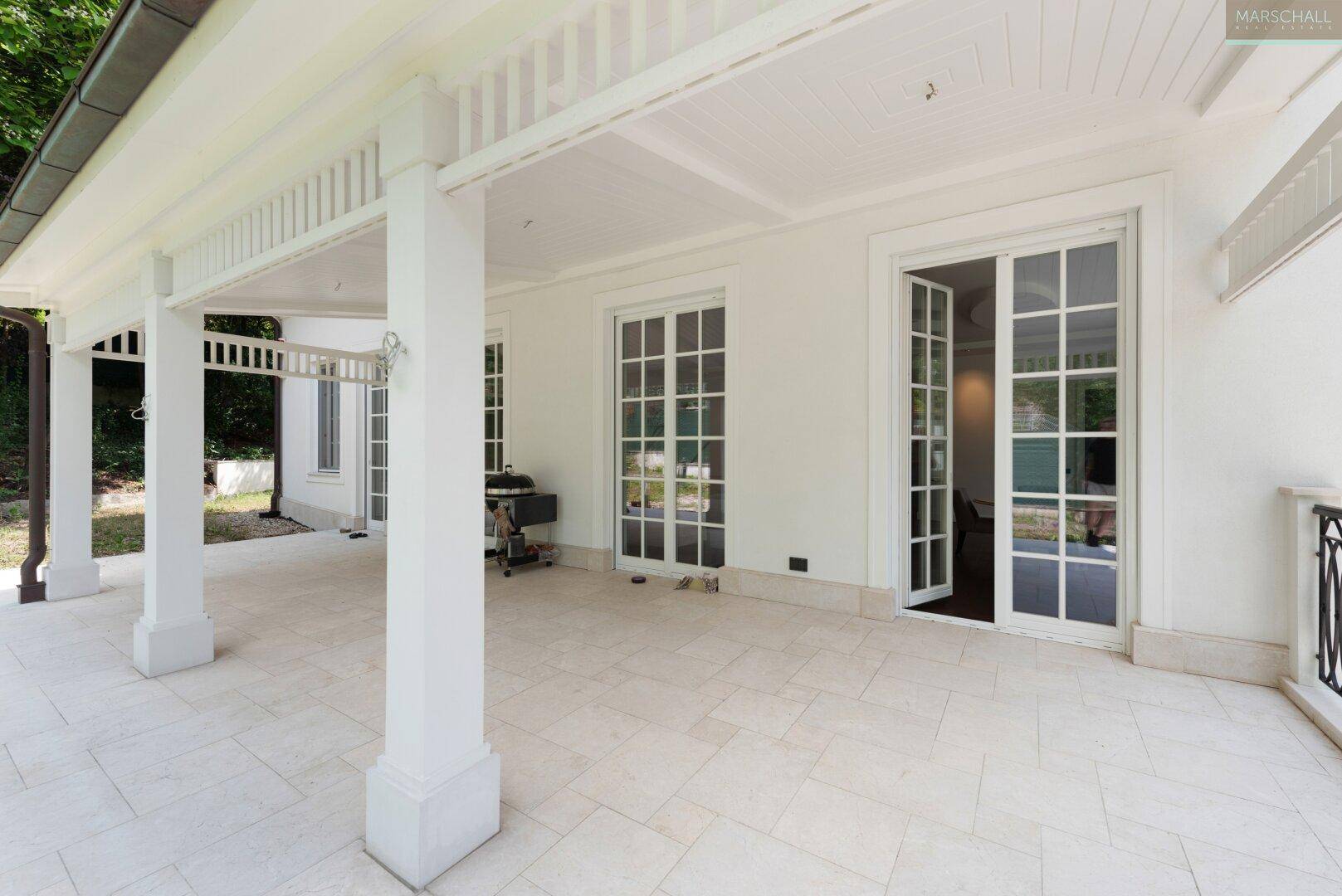 Überdachte Terrasse mit hellen Fliesen, weißen Säulen und Zugang zum Haus, ideal für Grillabende.