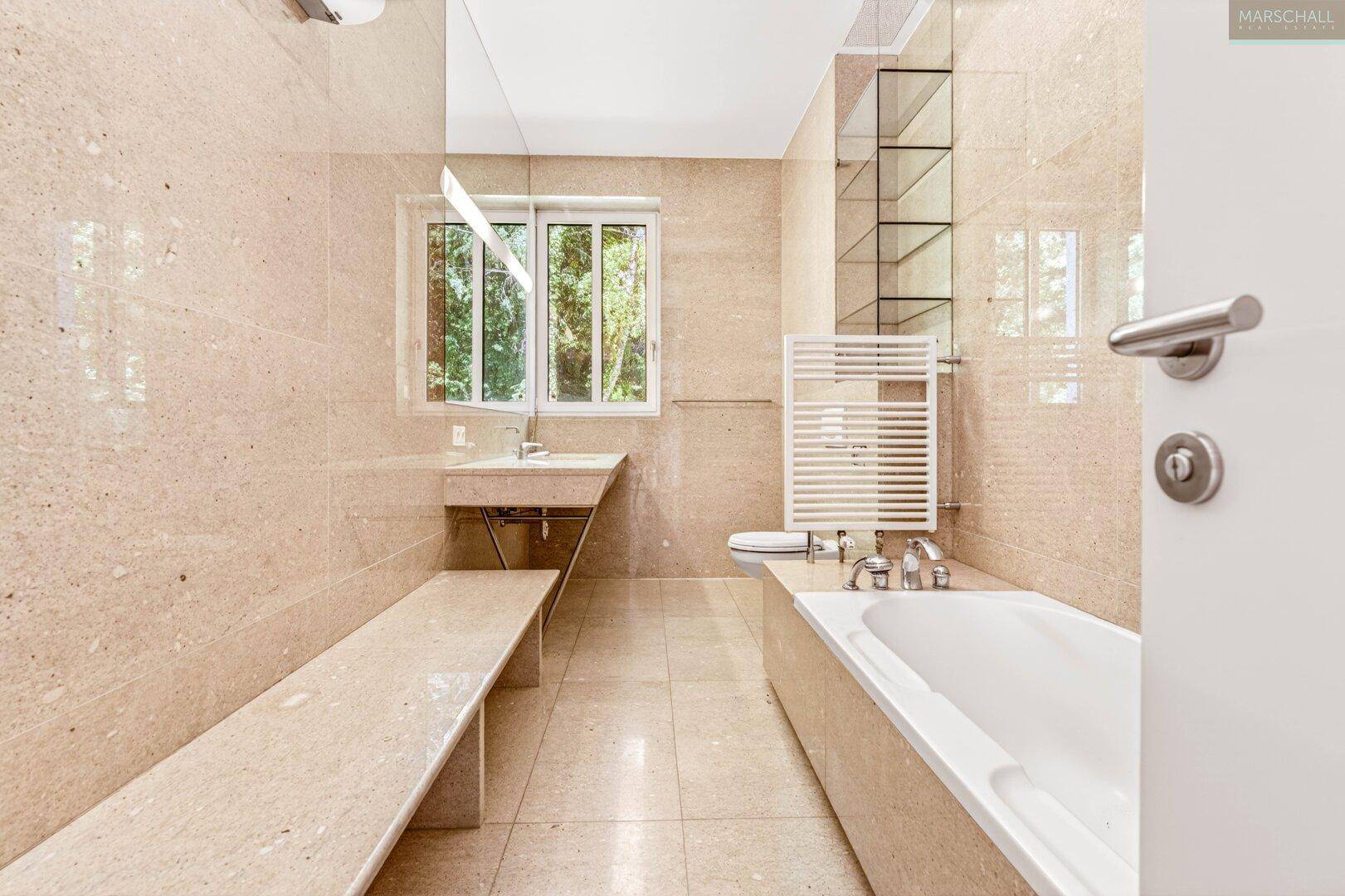 Helles Badezimmer mit Badewanne, beigefarbenen Fliesen und Fenster.