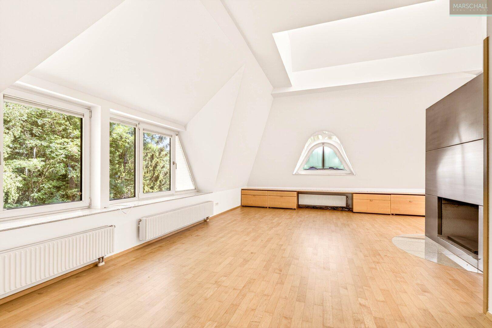Geräumiges Wohnzimmer mit Holzboden, Kamin und großen Fenstern mit Blick ins Grüne.