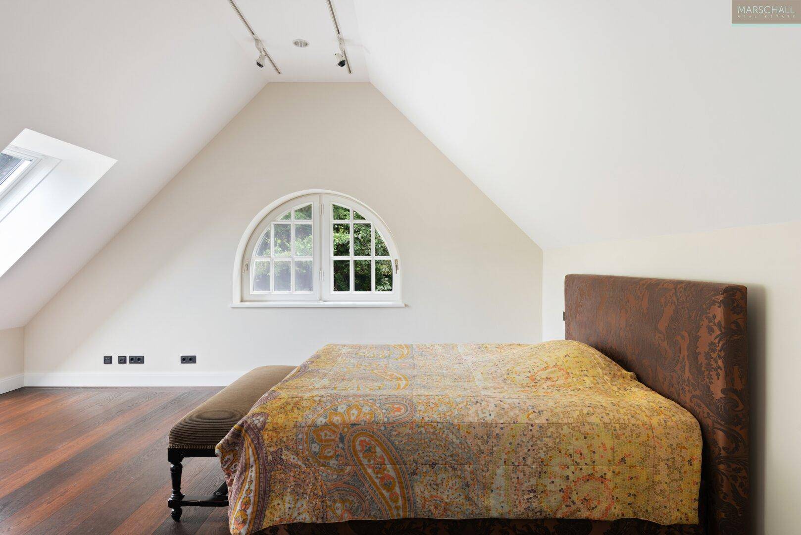 Schlafzimmer im Dachgeschoss mit schrägen Wänden, Holzboden und einem Rundbogenfenster.