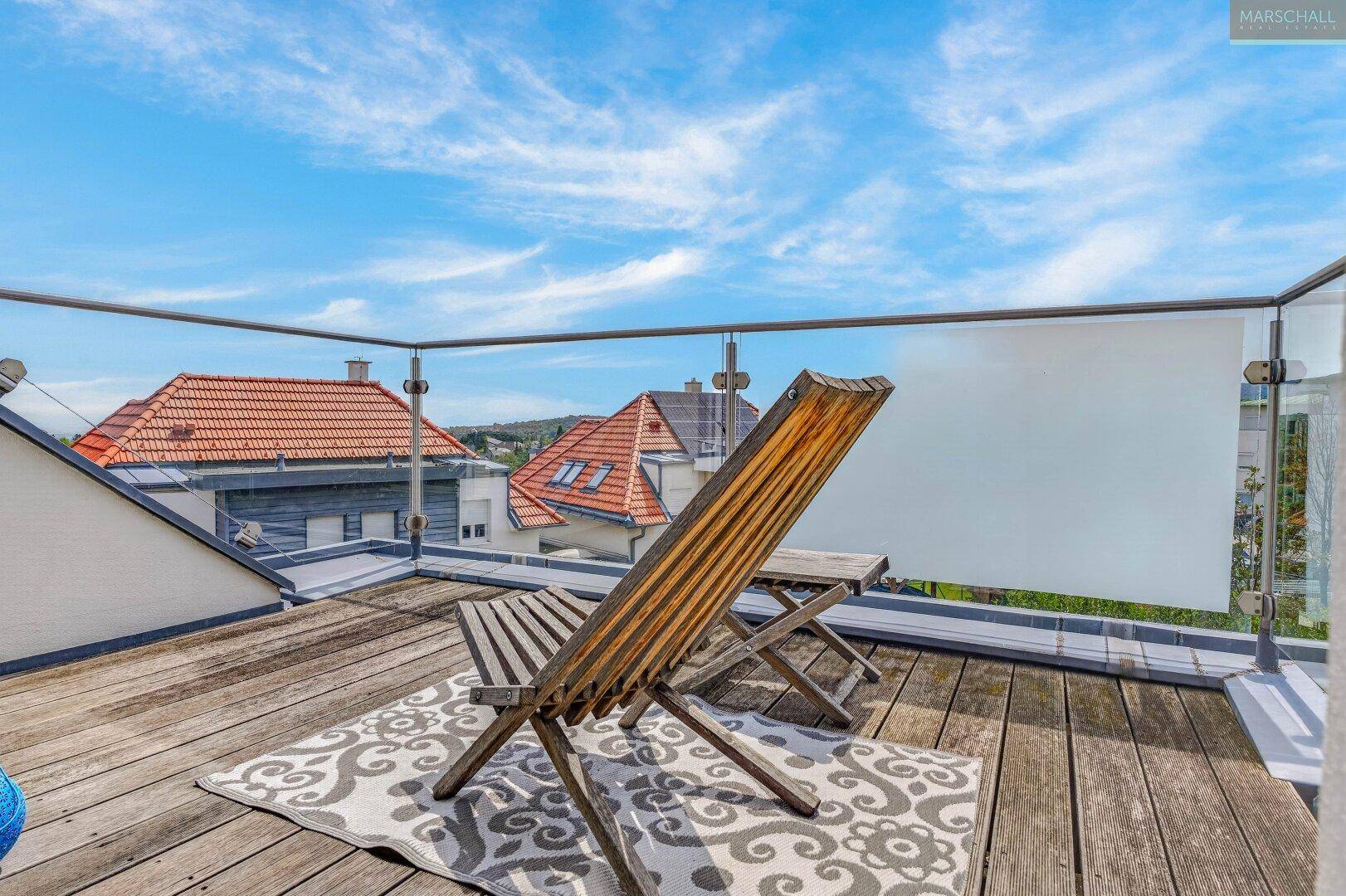 Sonniger Balkon mit Holzboden, Glasgeländer und Blick über die Dächer.