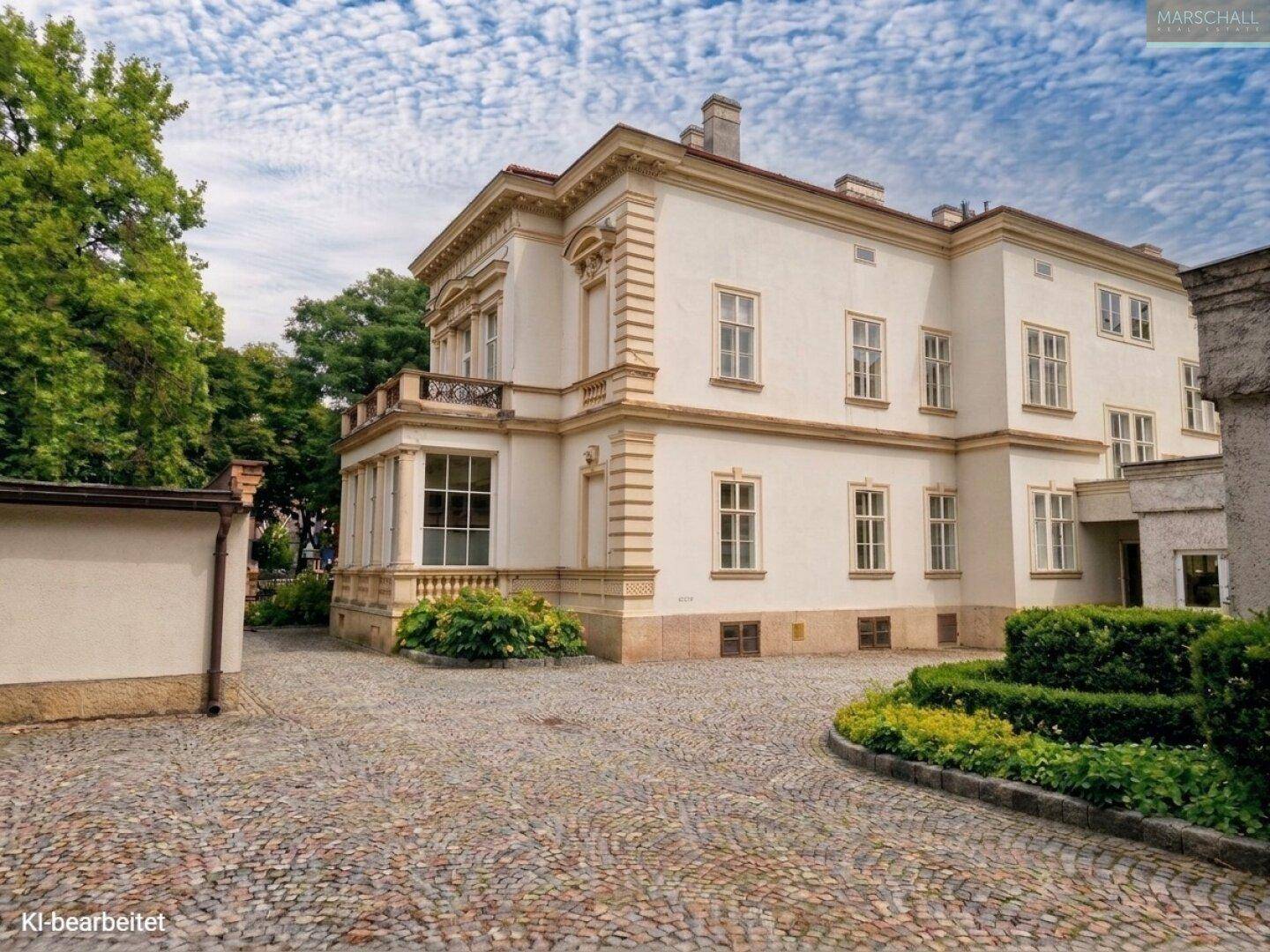Historisches Gebäude mit gepflastertem Vorplatz und gepflegten Hecken im Gartenbereich.