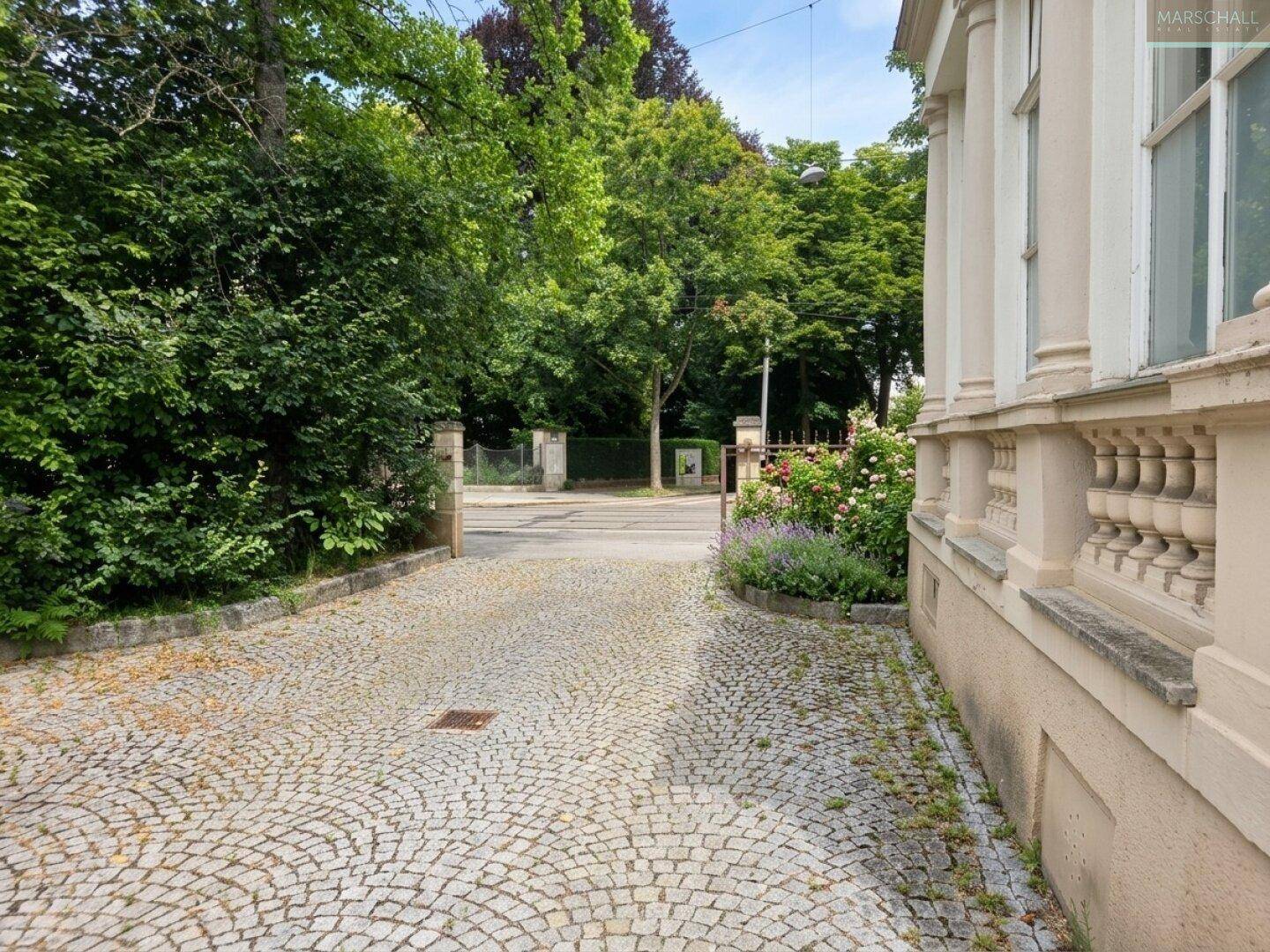 Gepflasterte Zufahrt zum Anwesen, umgeben von üppigem Grün und Bäumen.