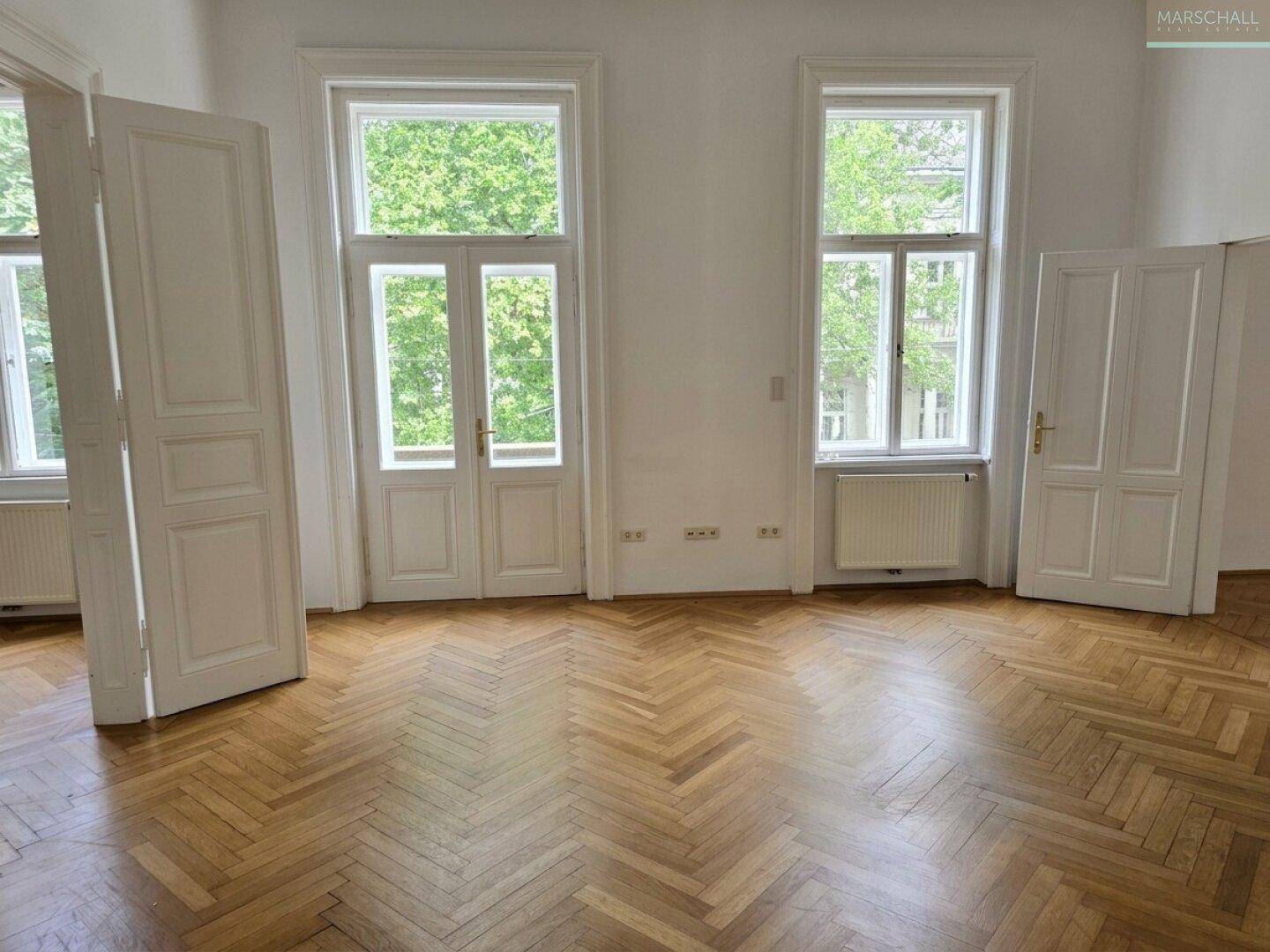 Großer, heller Raum mit Fischgrätparkett und mehreren Fenstern, die eine angenehme Atmosphäre schaffen.