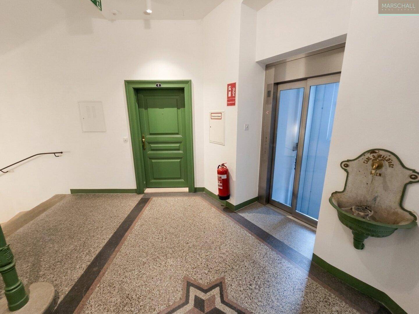 Eingangsbereich mit Terrazzoboden, Aufzug und einer historischen Wasserstelle an der Wand.