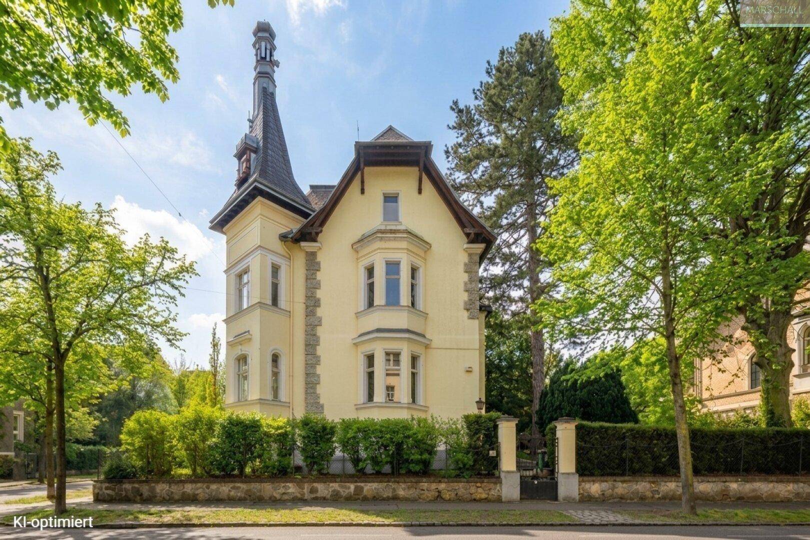 Elegante historische Villa mit Erker und hohem Turm, umgeben von altem Baumbestand.