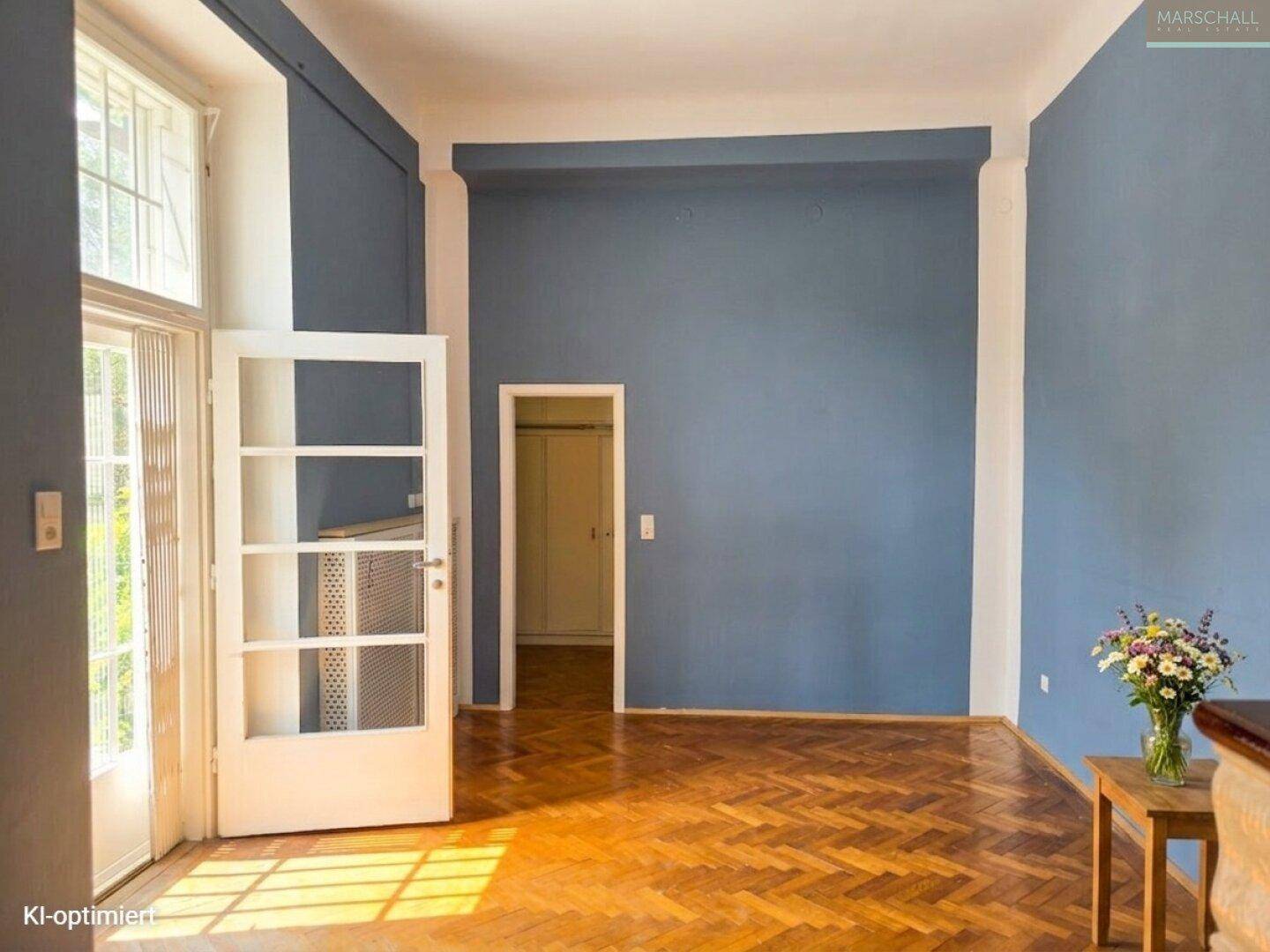 Geräumiges Zimmer mit blauen Wänden, Fischgrätparkett und Zugang zu einem Balkon.