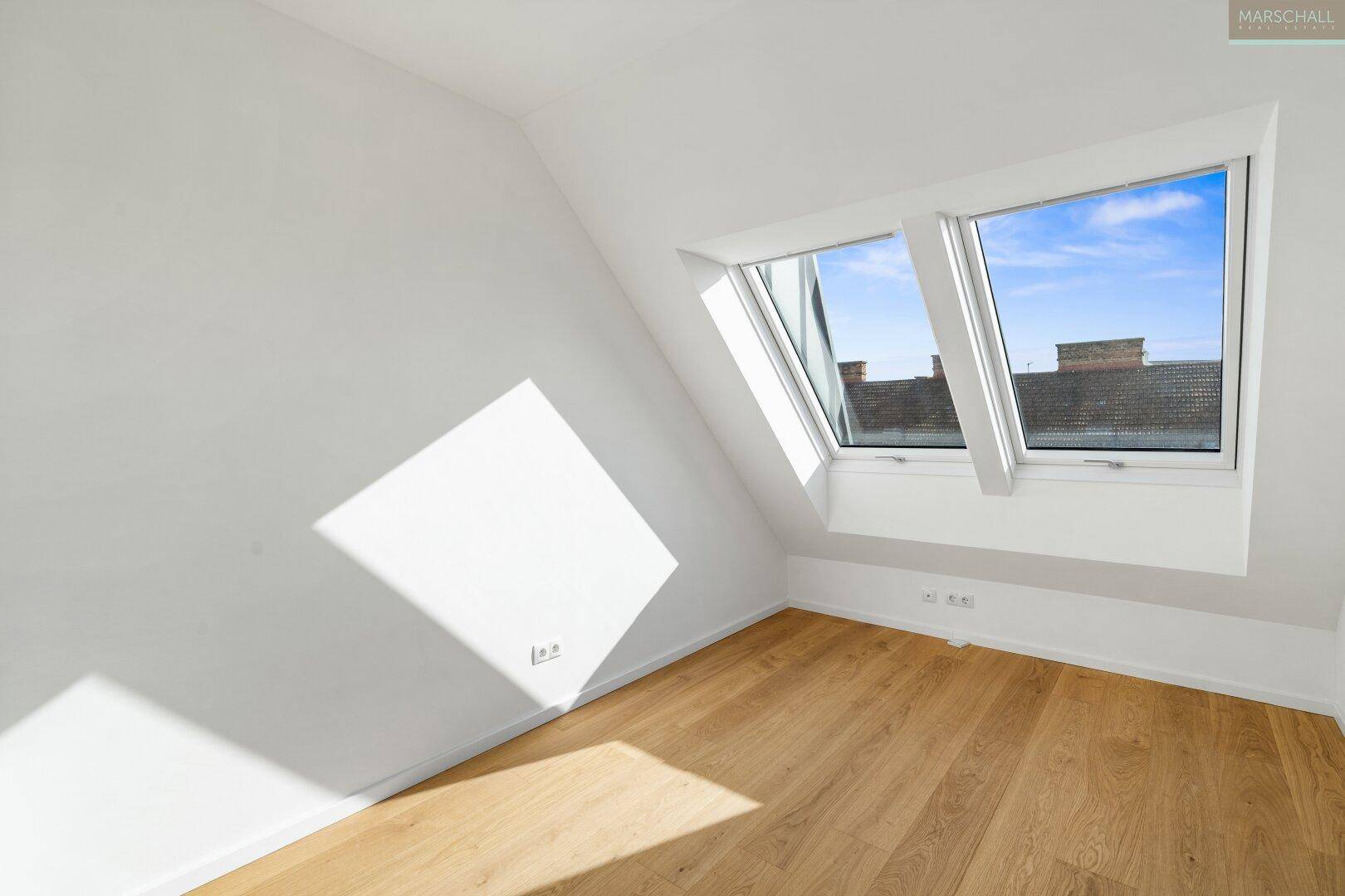 Heller Raum mit Holzboden und zwei schrägen Dachfenstern, die viel Sonnenlicht hereinlassen.