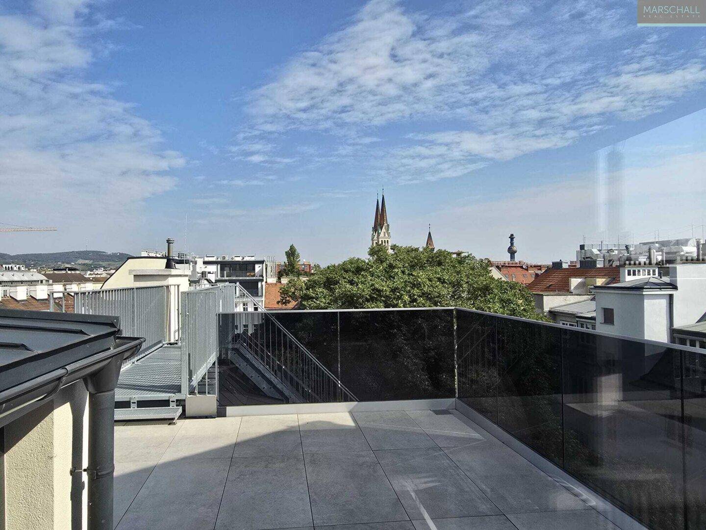 Geräumige Dachterrasse mit Glasgeländer und Zugang über eine Metalltreppe, bietet Stadtblick.