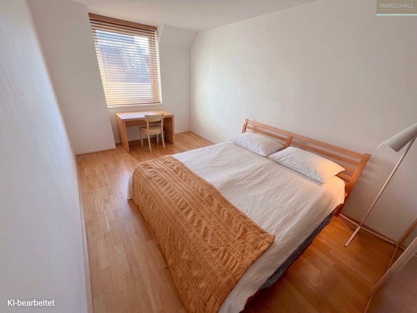 Helles Schlafzimmer mit Holzbett, Schreibtisch und Parkettboden unter einem Fenster mit Jalousien.