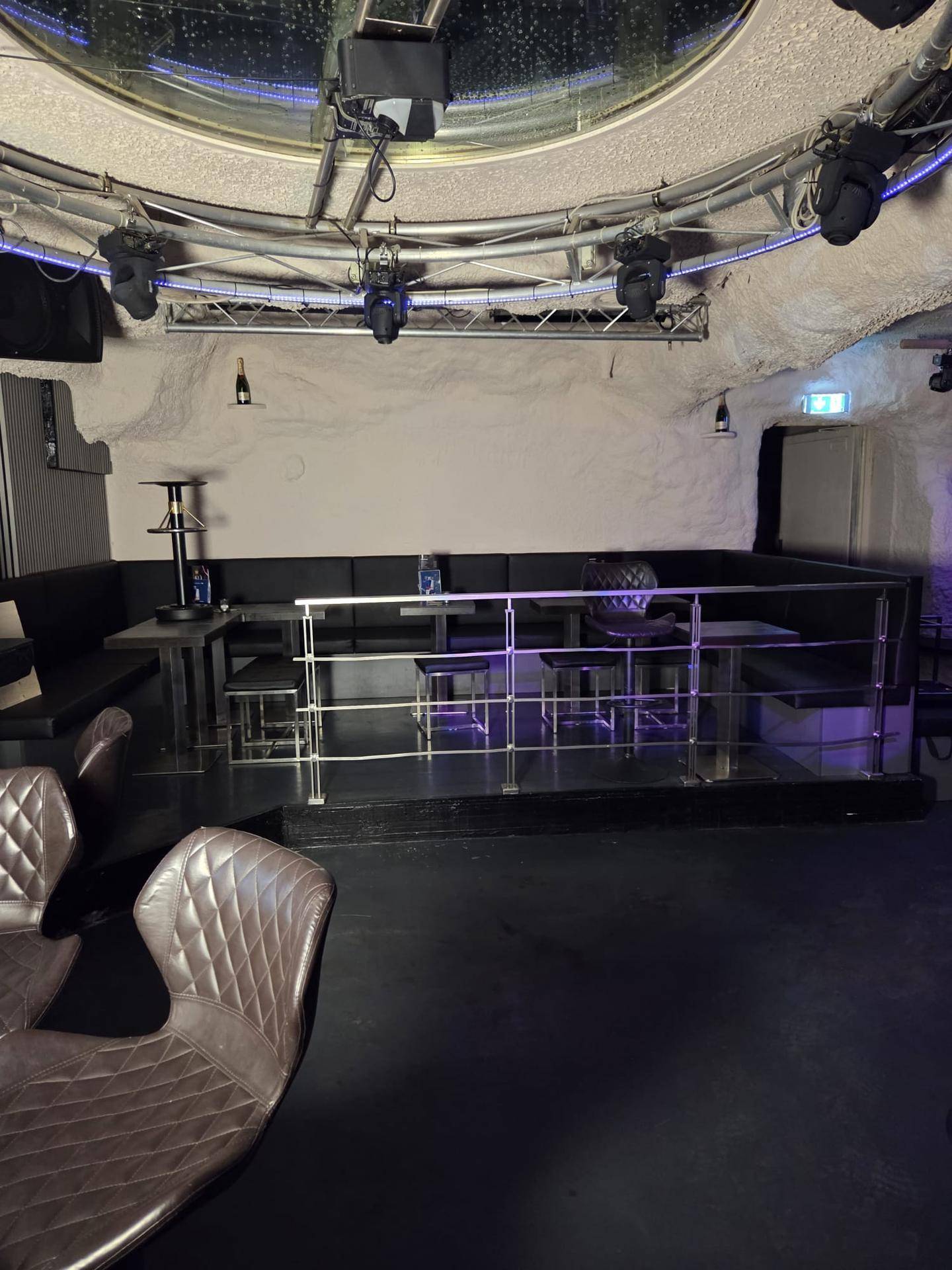 Sitzbereich einer Bar mit gepolsterten Bänken, Tischen und einer runden Deckenkonstruktion mit Beleuchtung.