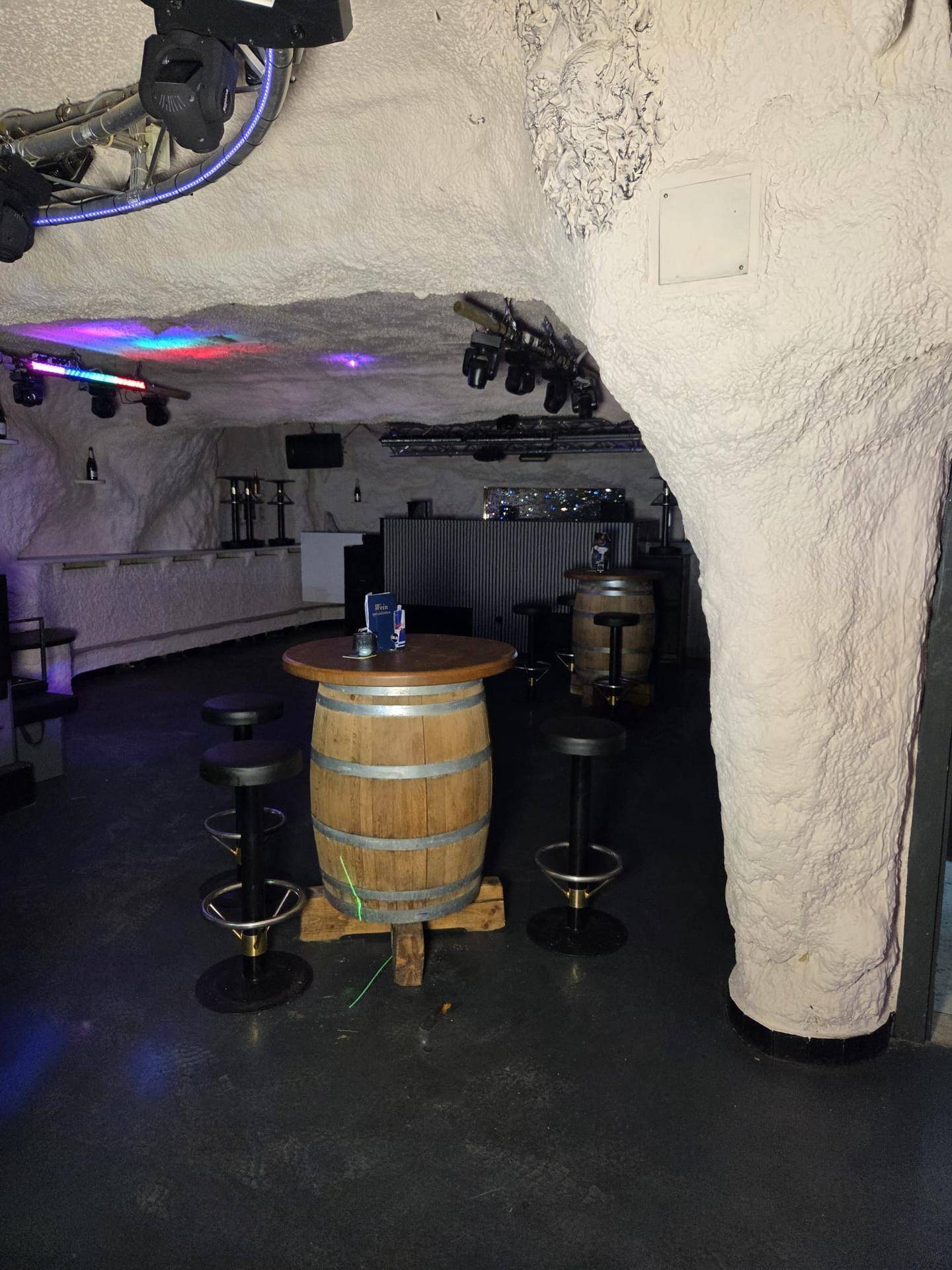 Innenansicht einer Bar mit rustikalem Fass-Tisch, Barhockern und stimmungsvoller Beleuchtung im Höhlen-Stil.