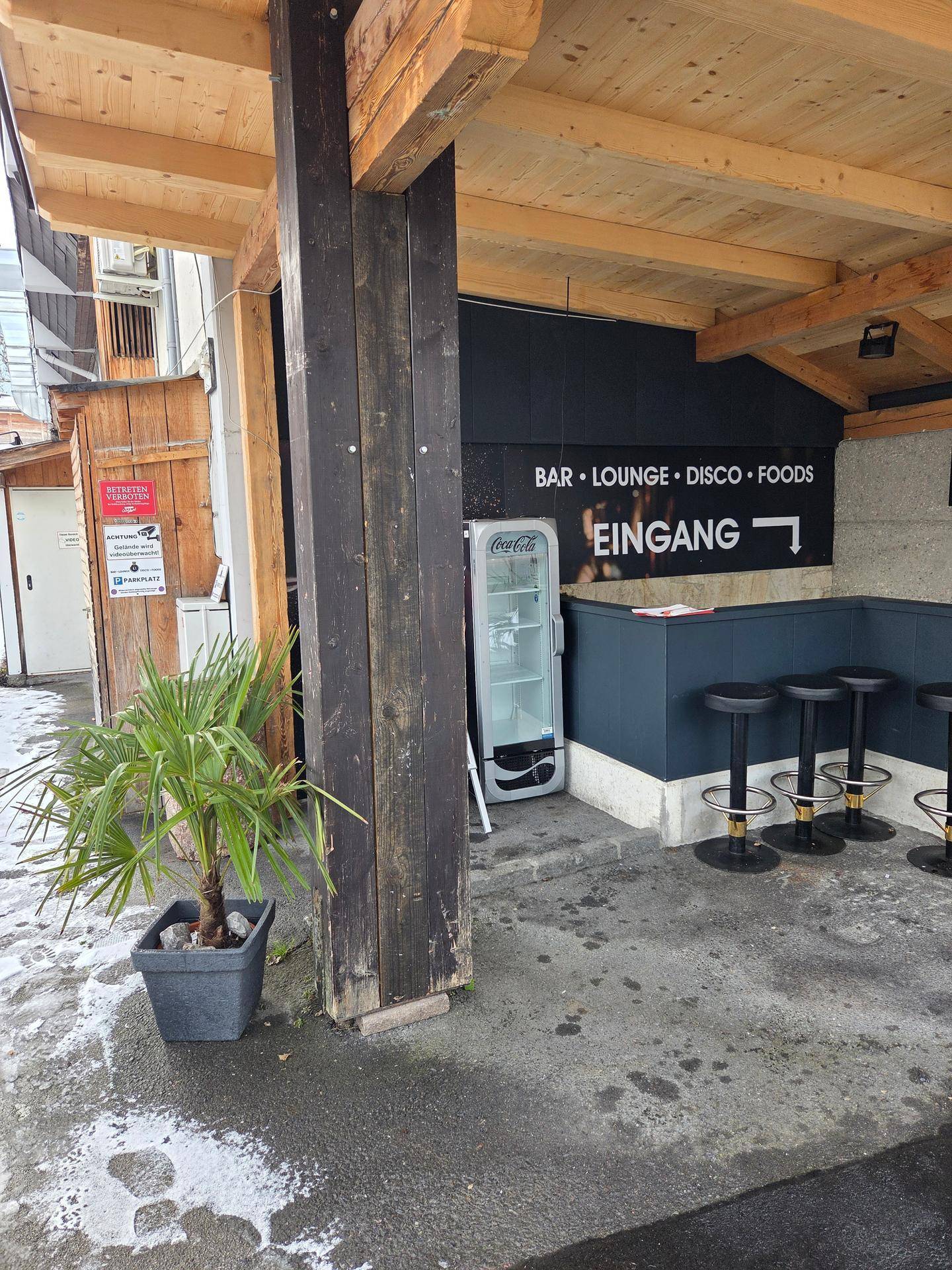 Eingangsbereich einer Bar mit Holzbalken, Beschilderung und einem Coca-Cola Kühlschrank im Freien.