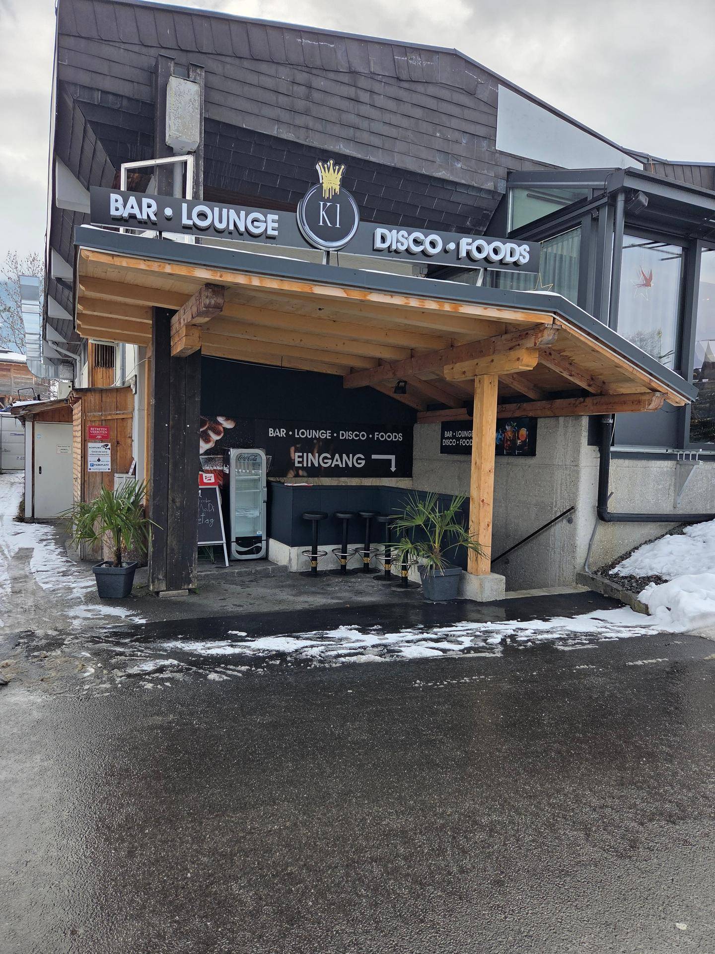 Gesamtansicht des Gebäudeeingangs einer Bar mit Holzdach, Beschilderung und winterlicher Umgebung.