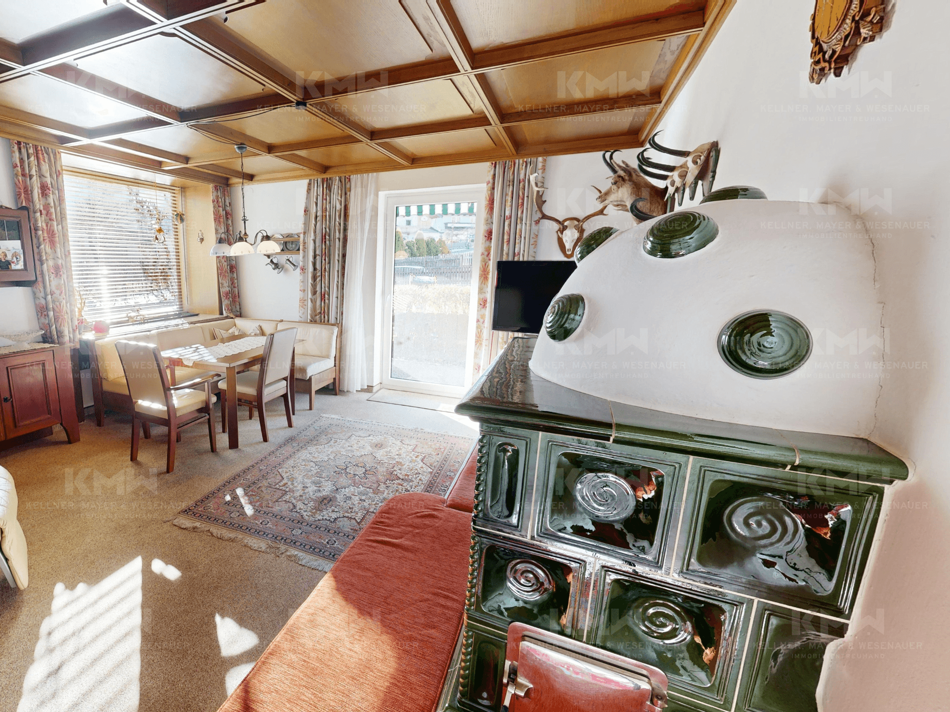 Gemütlicher Wohn-Essbereich mit traditionellem Kachelofen, Holzbalkendecke und Teppichboden.