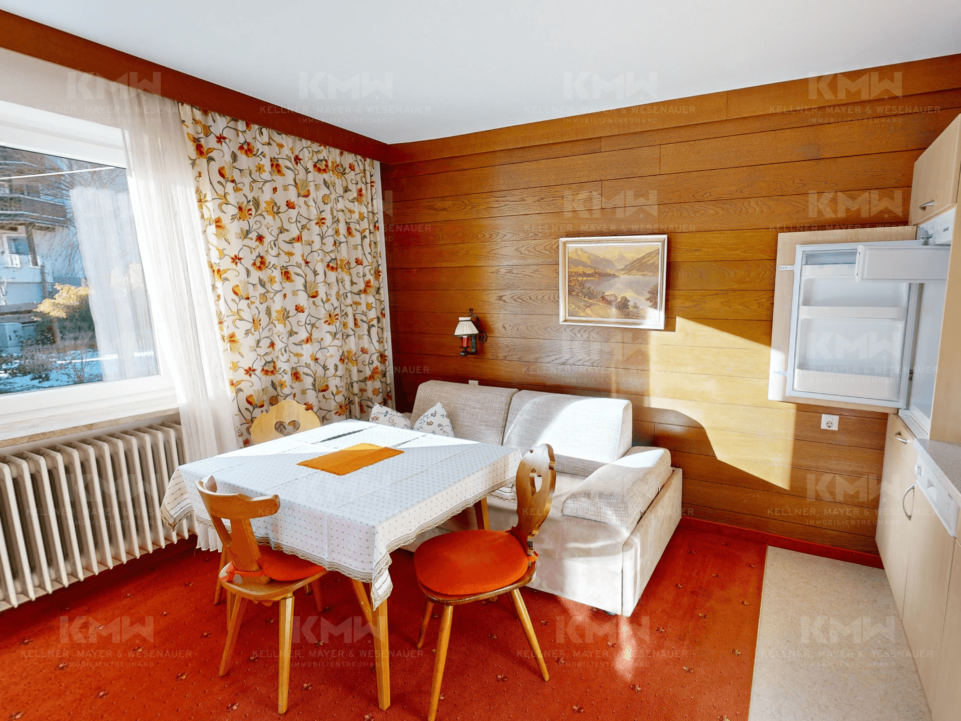 Wohn- und Essbereich mit Holzvertäfelung, Teppichboden und floralen Vorhängen.