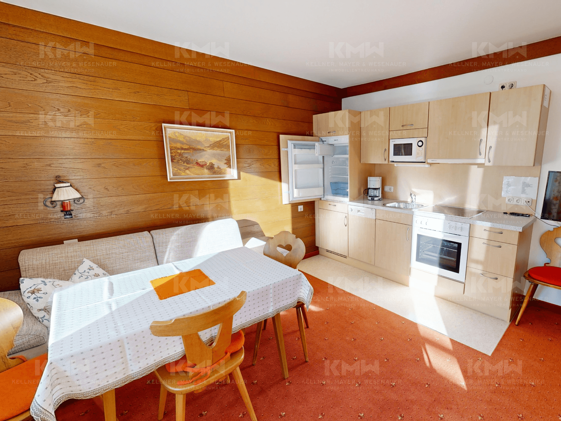 Kleine Küchenzeile mit integrierten Geräten, Esstisch und Holzvertäfelung im Wohnbereich.