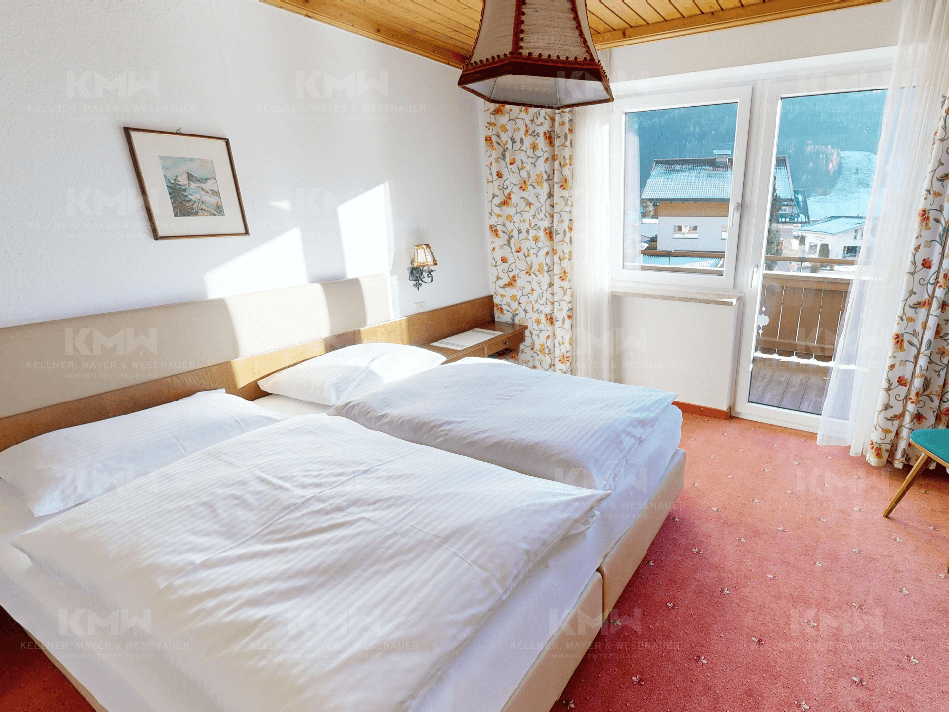 Helles Schlafzimmer mit Doppelbett, Holzbalkendecke und Zugang zu einem kleinen Balkon mit Bergblick.