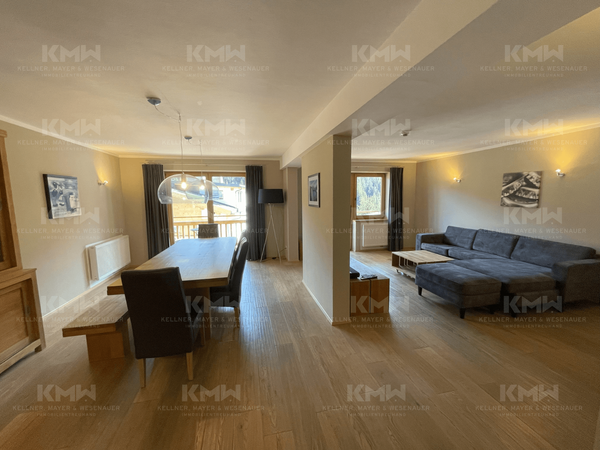 Weitläufiger Wohn-Essbereich mit großem Sofa, Holzboden und viel Tageslicht durch die Fenster.