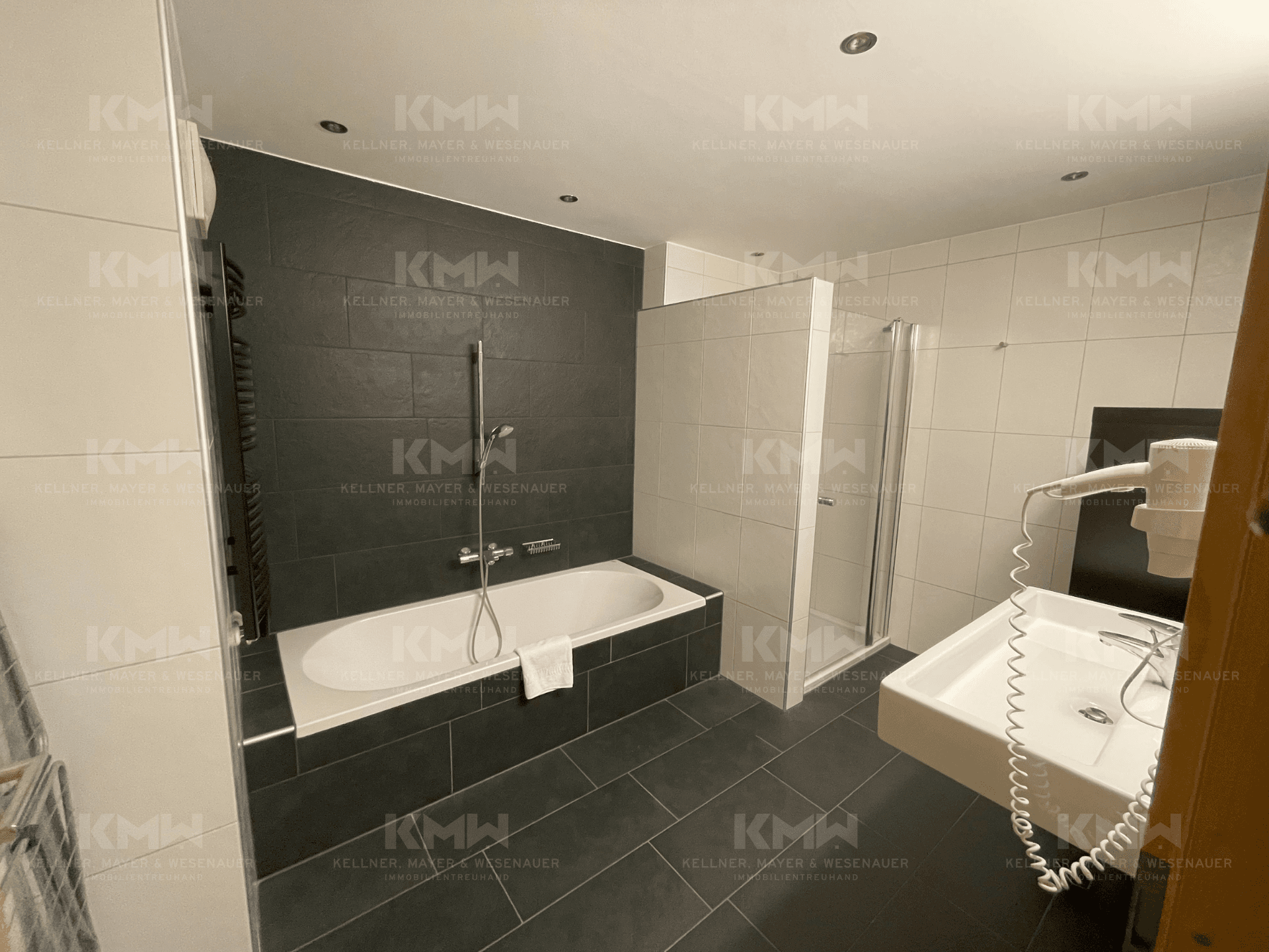 Modernes Badezimmer mit Badewanne, Dusche und dunklen Fliesen an der Wand und auf dem Boden.