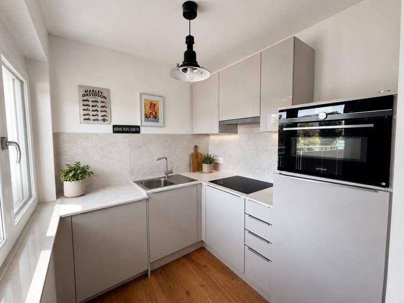 Moderne Küche mit hellgrauen Schränken, Holzboden und integrierten Geräten, gut beleuchtet durch ein Fenster.