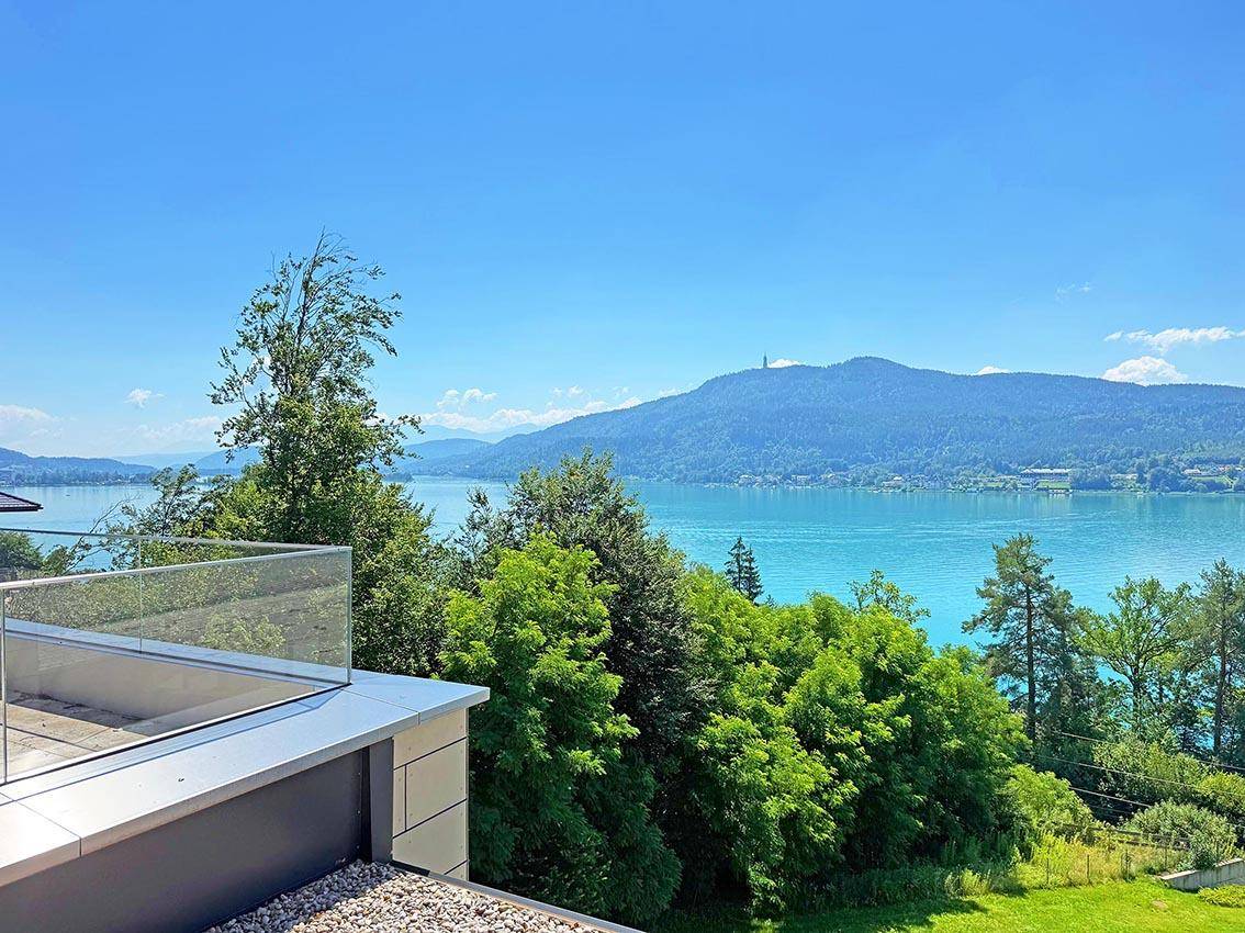 Panoramablick von einer Terrasse auf den See, bewaldete Hügel und ferne Berge unter blauem Himmel.
