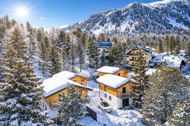 Winterliche Luftaufnahme von Chalets in einer verschneiten Berglandschaft unter strahlendem Sonnenschein.