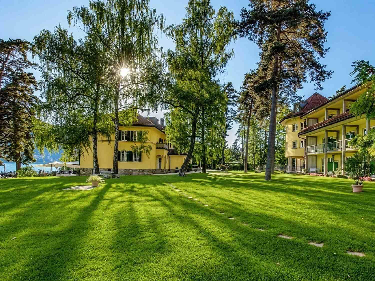 Weitläufige, sonnige Rasenfläche mit hohen Bäumen und Blick auf die gelben Gebäude des Anwesens.